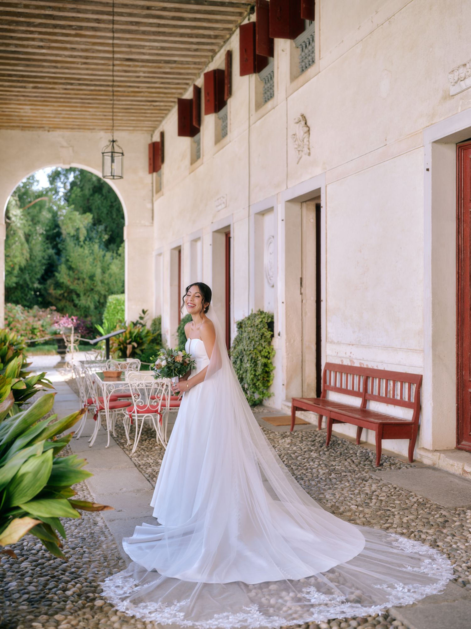 A flowing white wedding dress with a long train is displayed along a rustic covered walkway with stone columns.