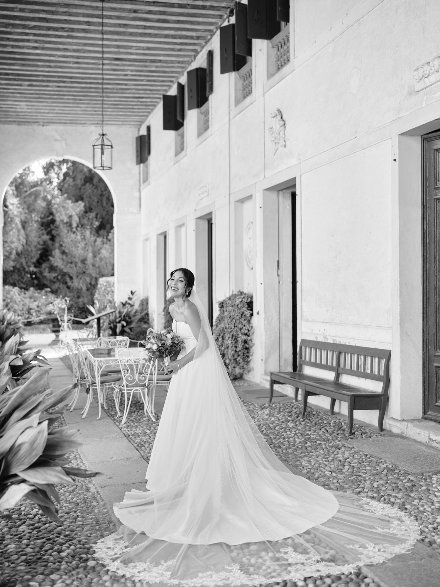 A flowing white wedding dress twirls gracefully across a stone porch with classic white columns in a black and white photo.