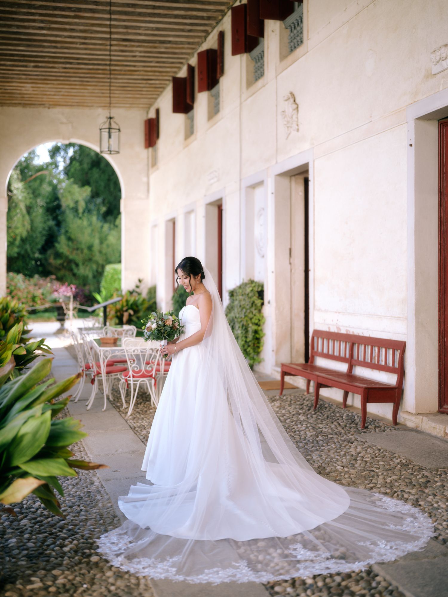 A bride in a flowing white wedding gown with a long veil poses along a rustic mission-style corridor with arches.