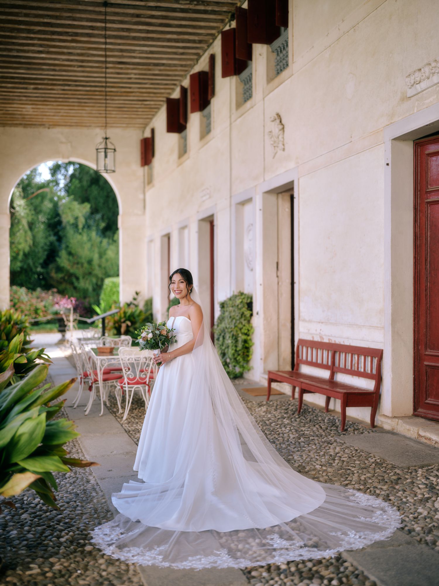 A wedding dress with a long flowing train photographed along a rustic Spanish colonial style covered walkway.