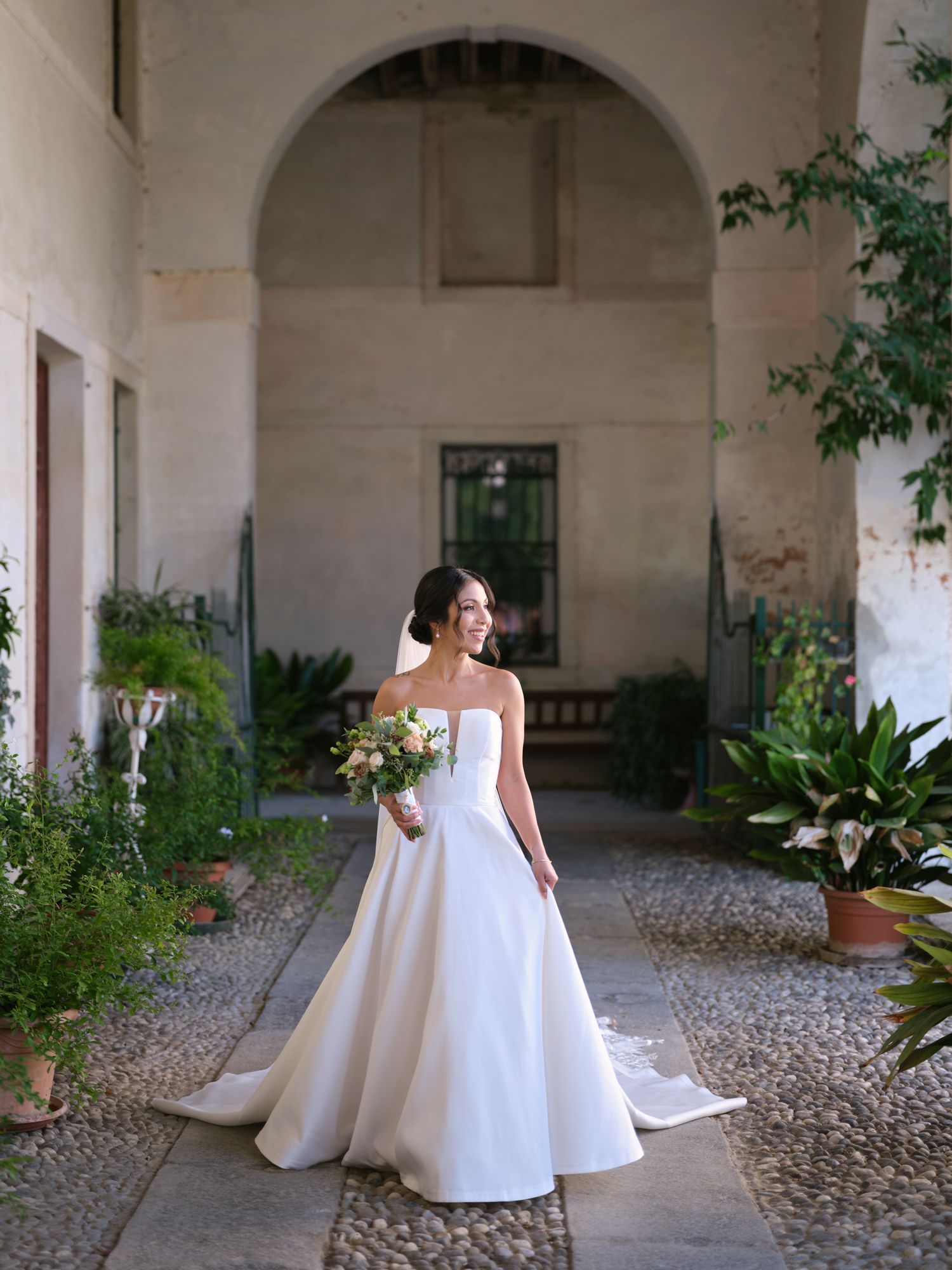 A bride in a flowing white ballgown stands under a grand stone archway in a historic courtyard.