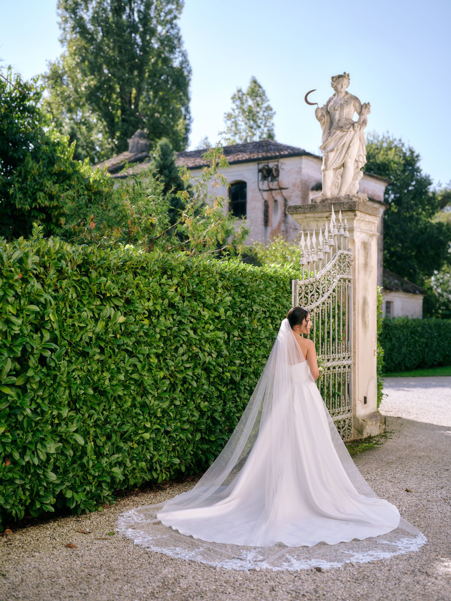A bride in a flowing white gown poses elegantly by ornate garden gates and manicured hedges at a grand estate.
