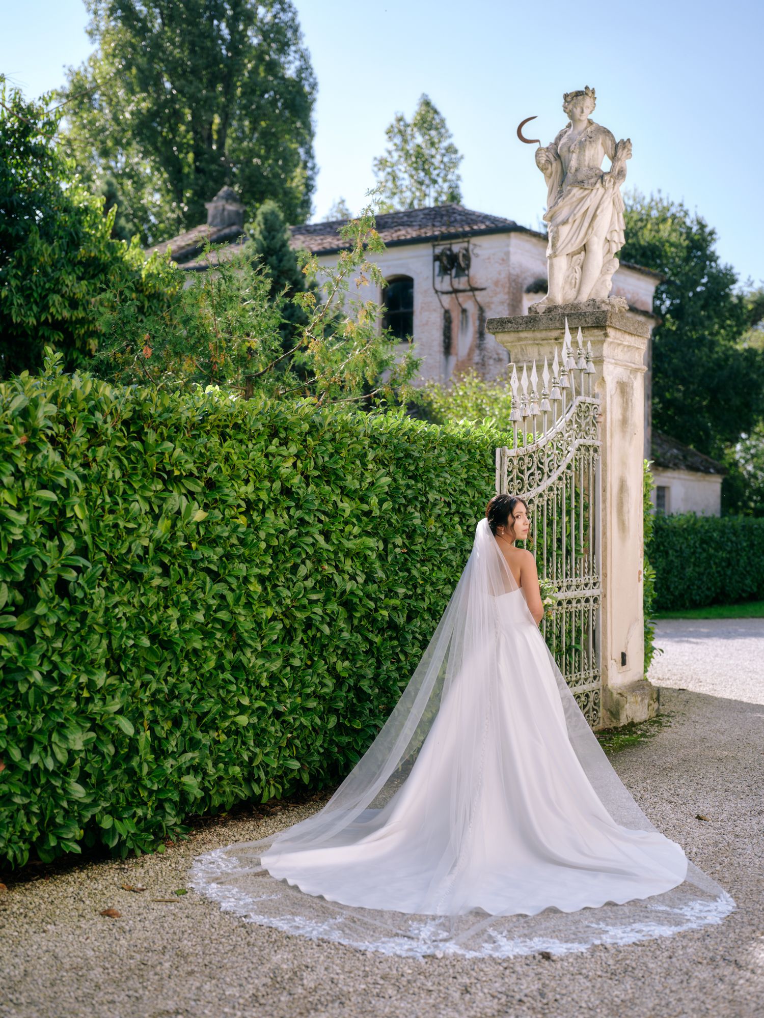 A bride in a flowing white ballgown poses gracefully by an ornate garden gate at a luxurious estate.