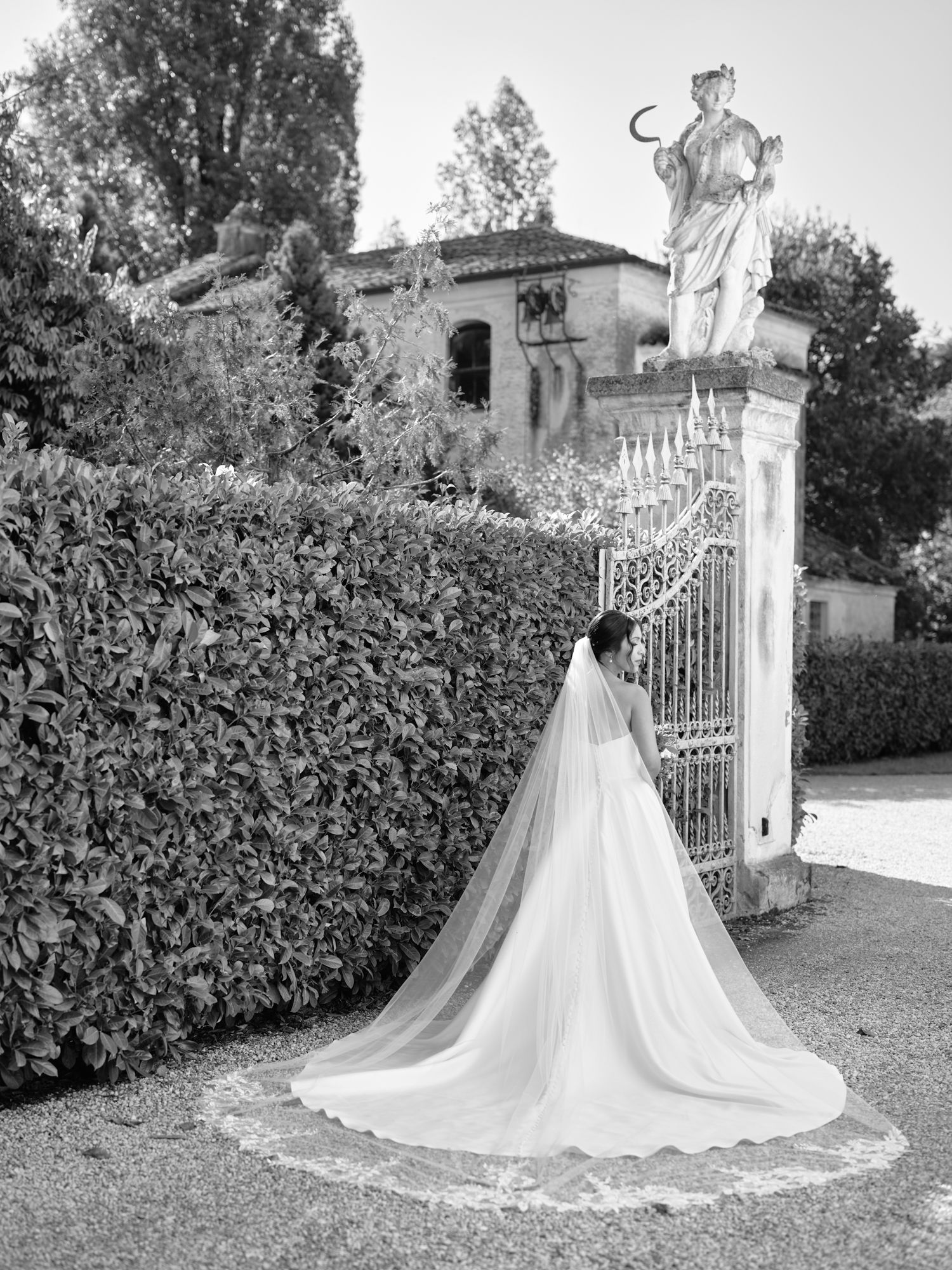 A flowing white wedding dress with a long cathedral train displayed against an ornate garden gate and hedges.