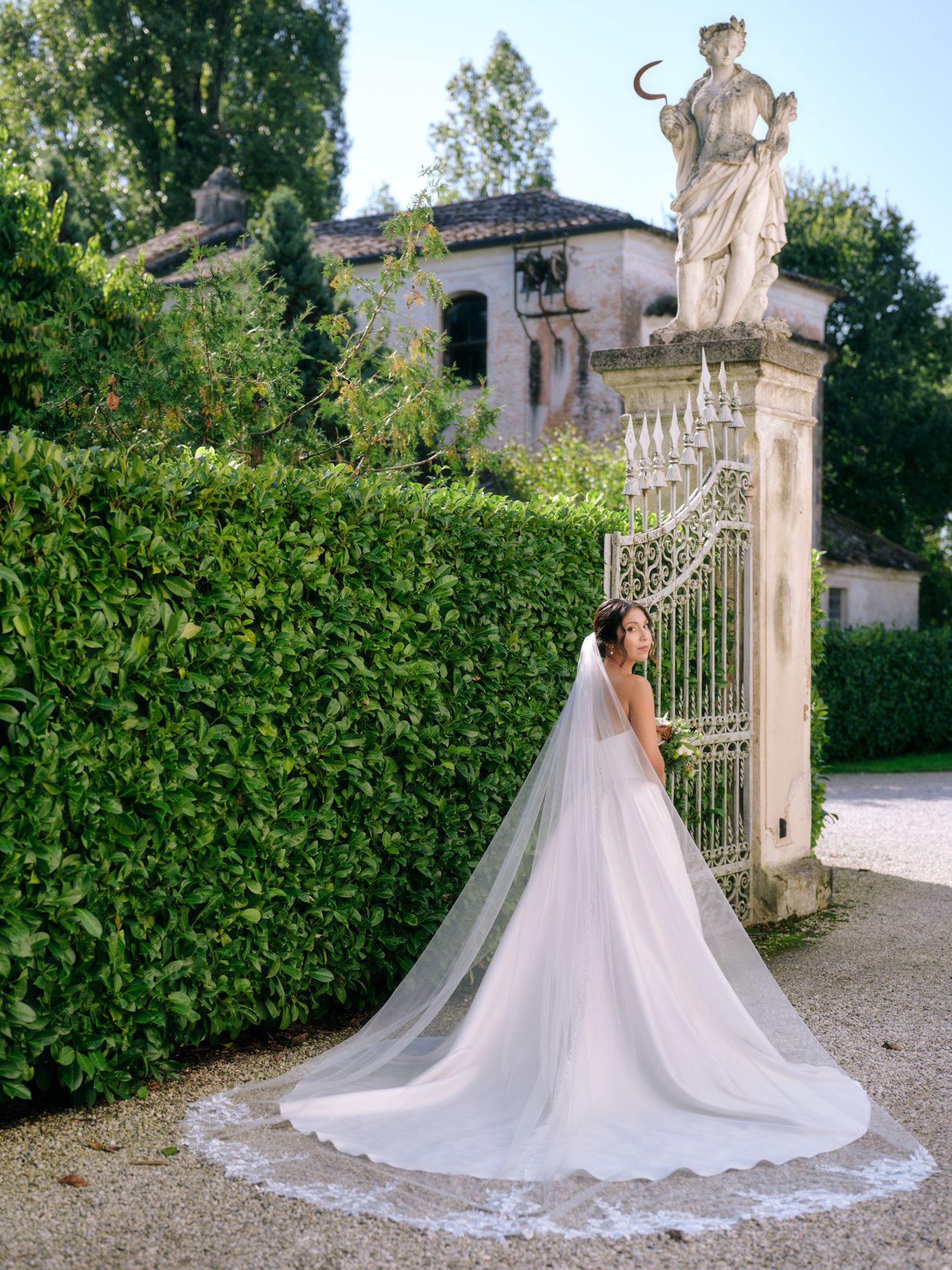 A bride in a flowing white gown with long cathedral veil poses by ornate garden gates and classical statuary.