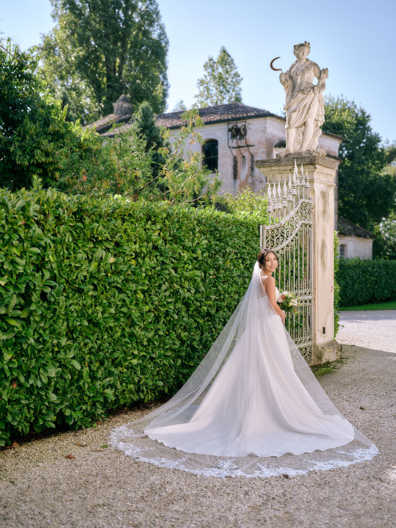 A bride in a flowing white gown with a long train poses elegantly in front of an ornate garden gate and hedge wall.