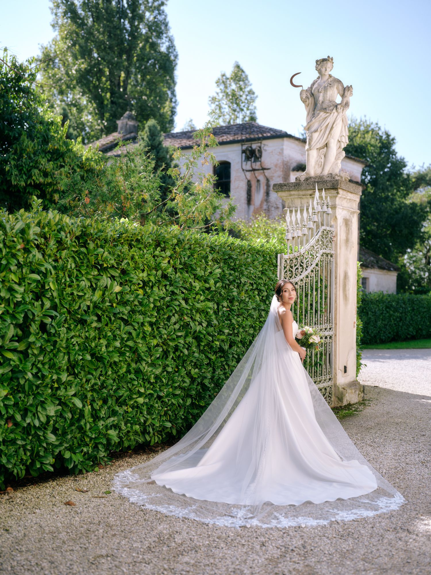 A bride in a flowing white gown with a dramatic cathedral-length veil poses elegantly by an ornate garden gate.
