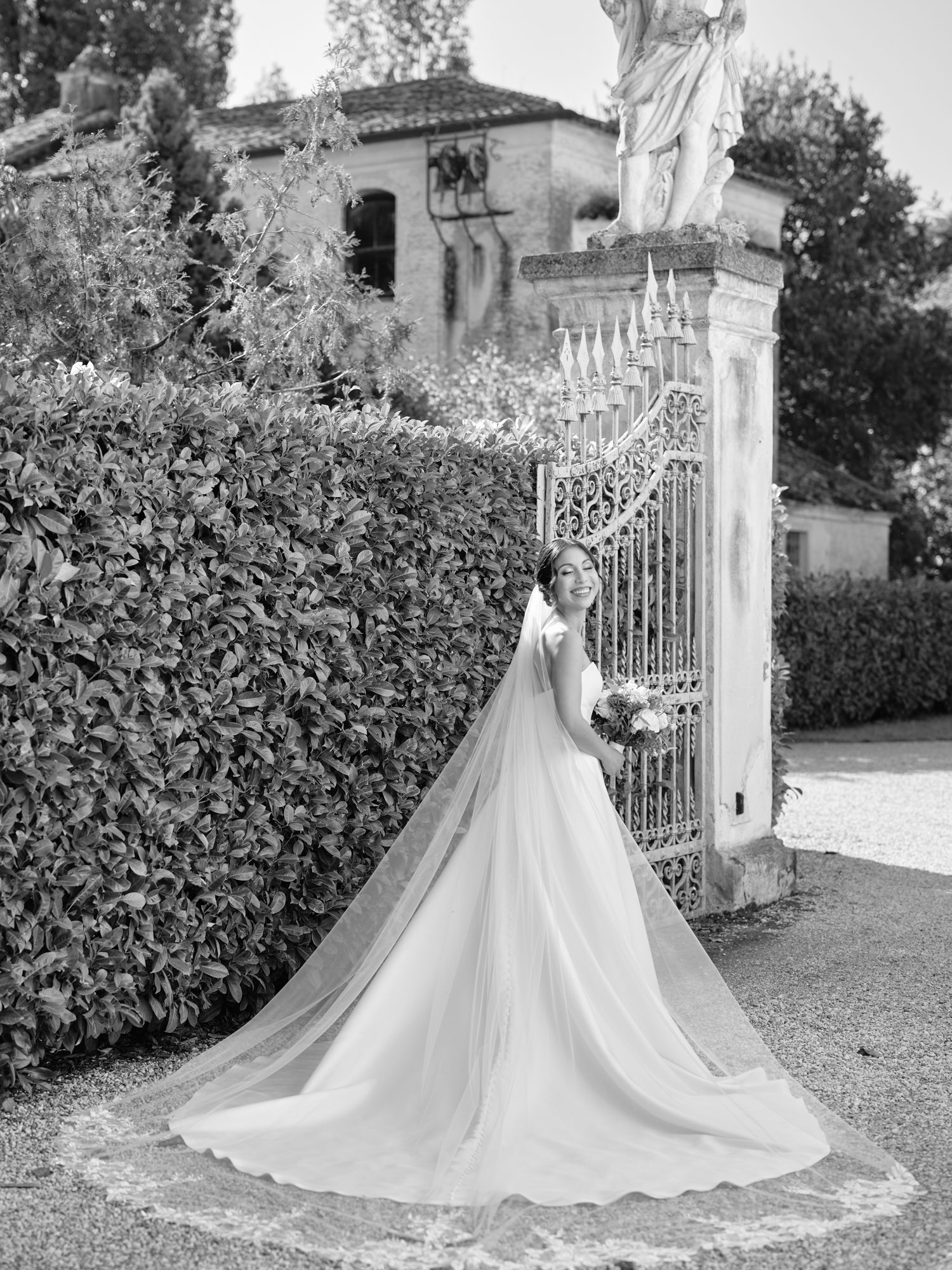 Black and white photo of a wedding dress with long flowing train by ornate garden gates and statue at elegant estate.
