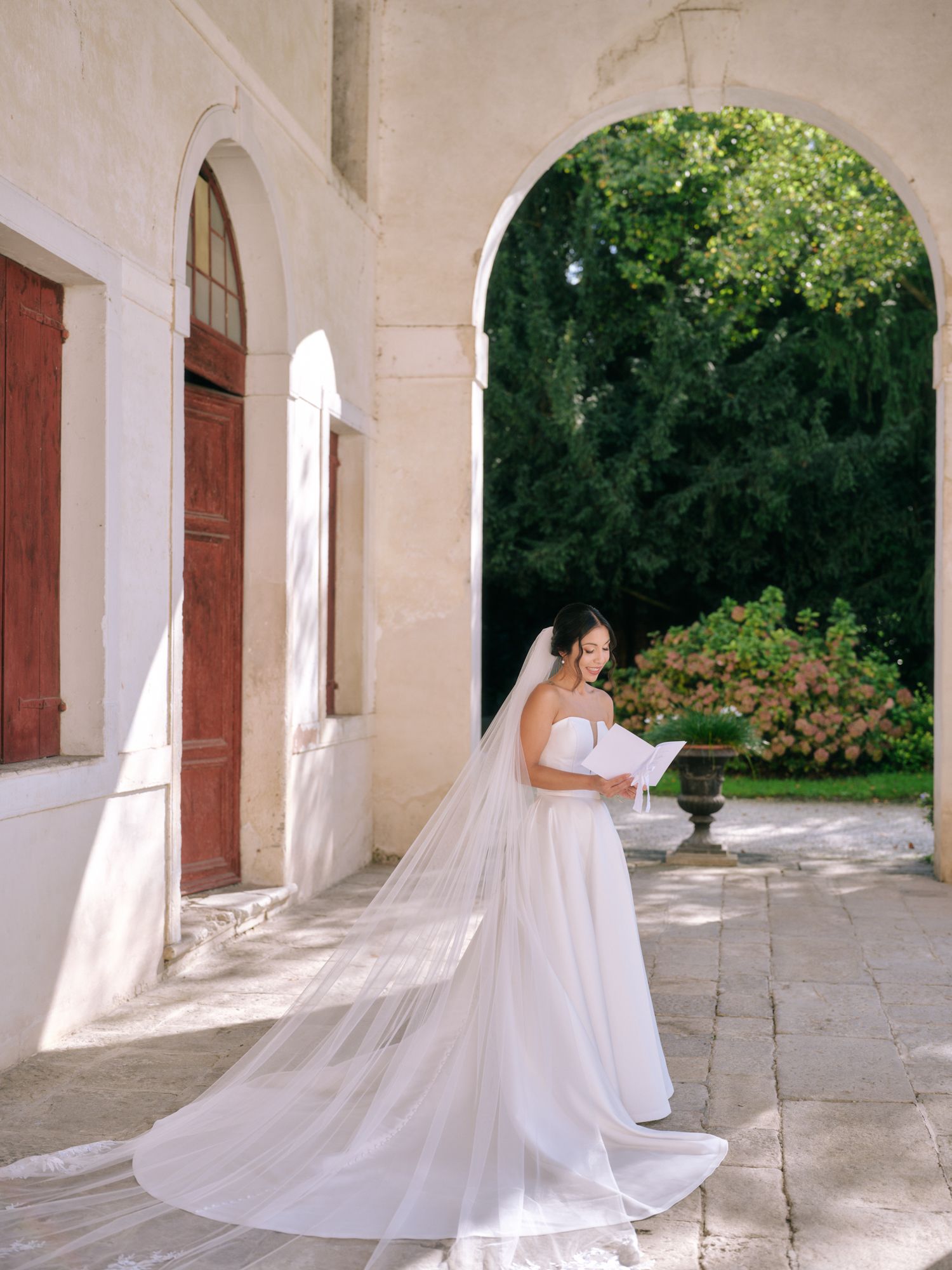 A flowing white wedding dress with long train creates an elegant silhouette against a stone colonnade walkway in natural light.