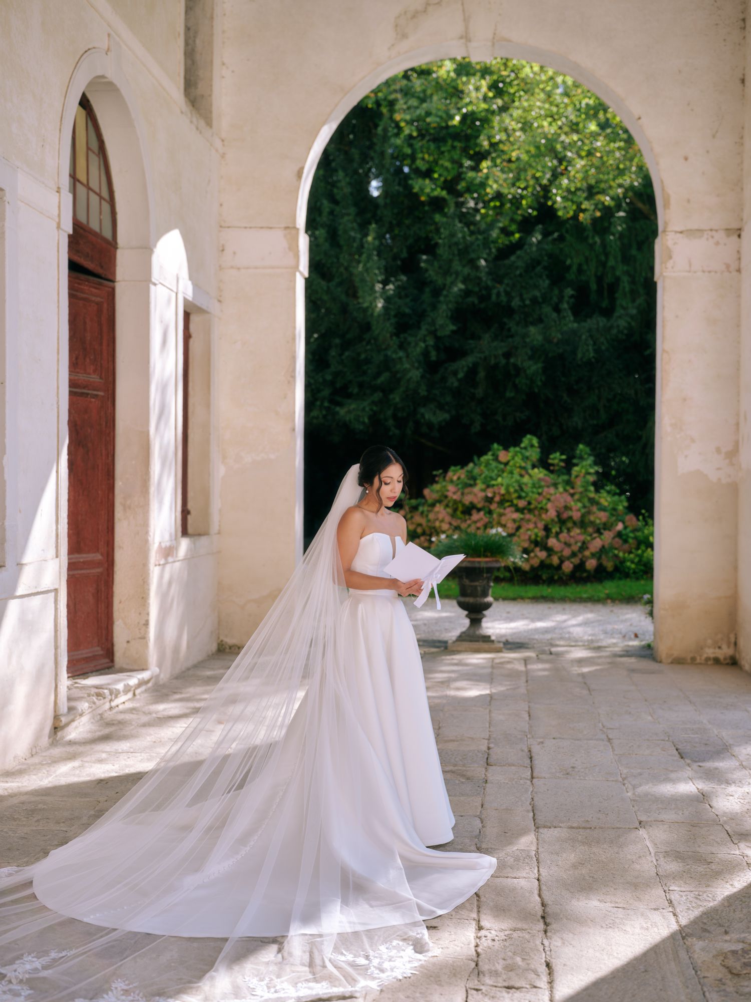 A flowing white wedding veil trails gracefully through a series of stone archways in an elegant architectural setting.
