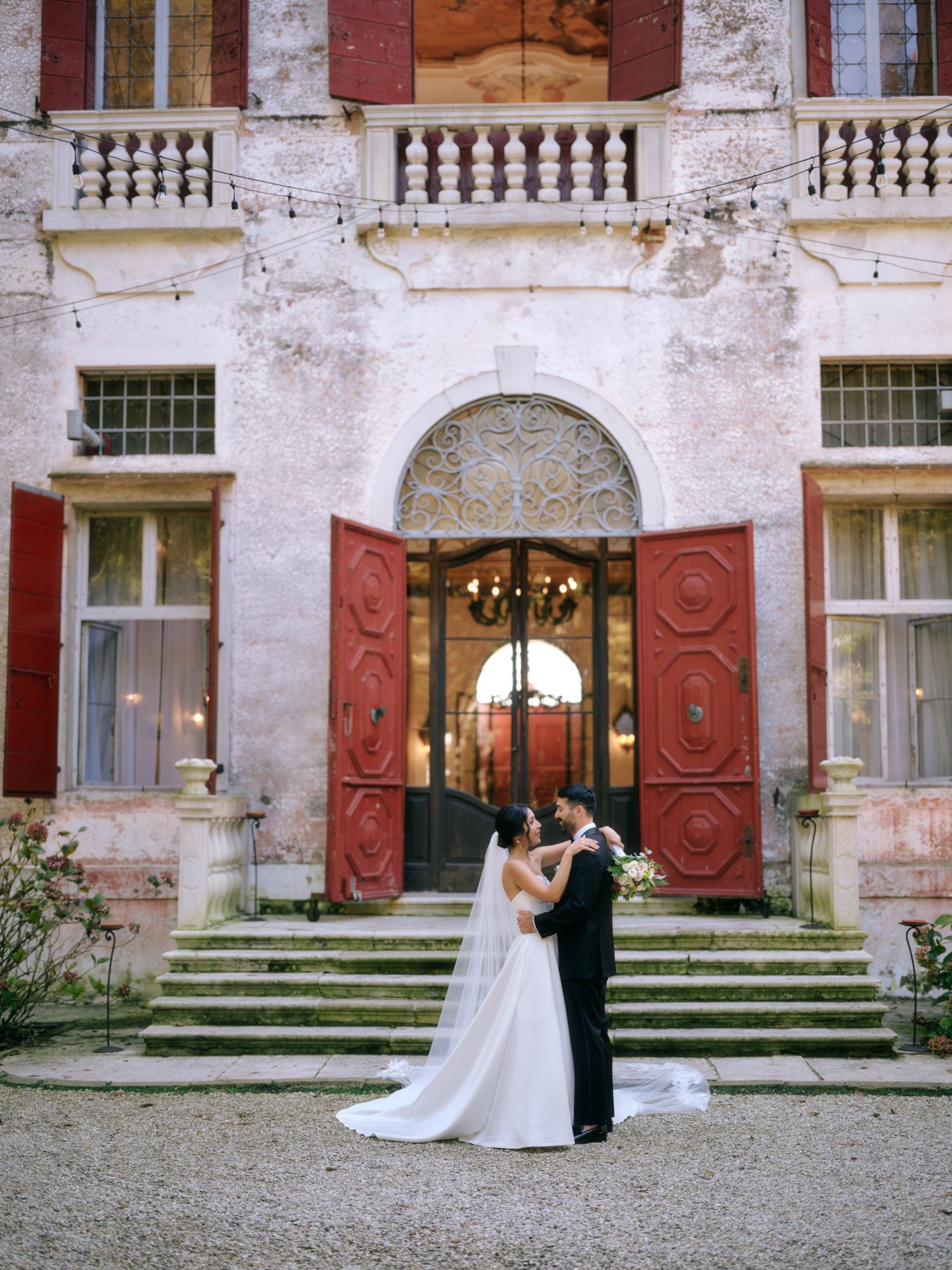 A wedding couple dances together on stone steps in front of an elegant building with red shutters and doors.