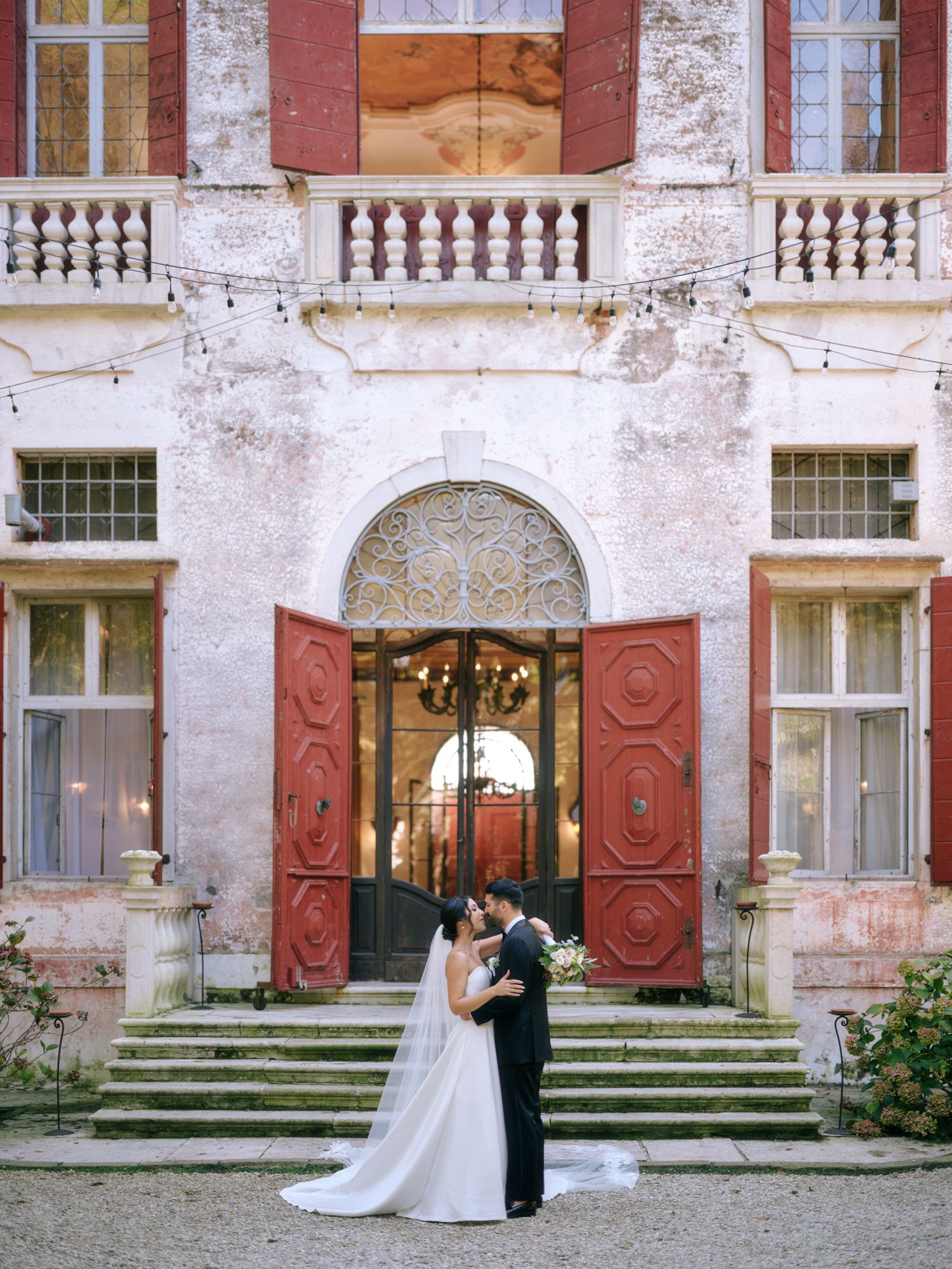 A romantic couple embraces on the steps of a historic mansion with weathered red and white walls and ornate balustrades.