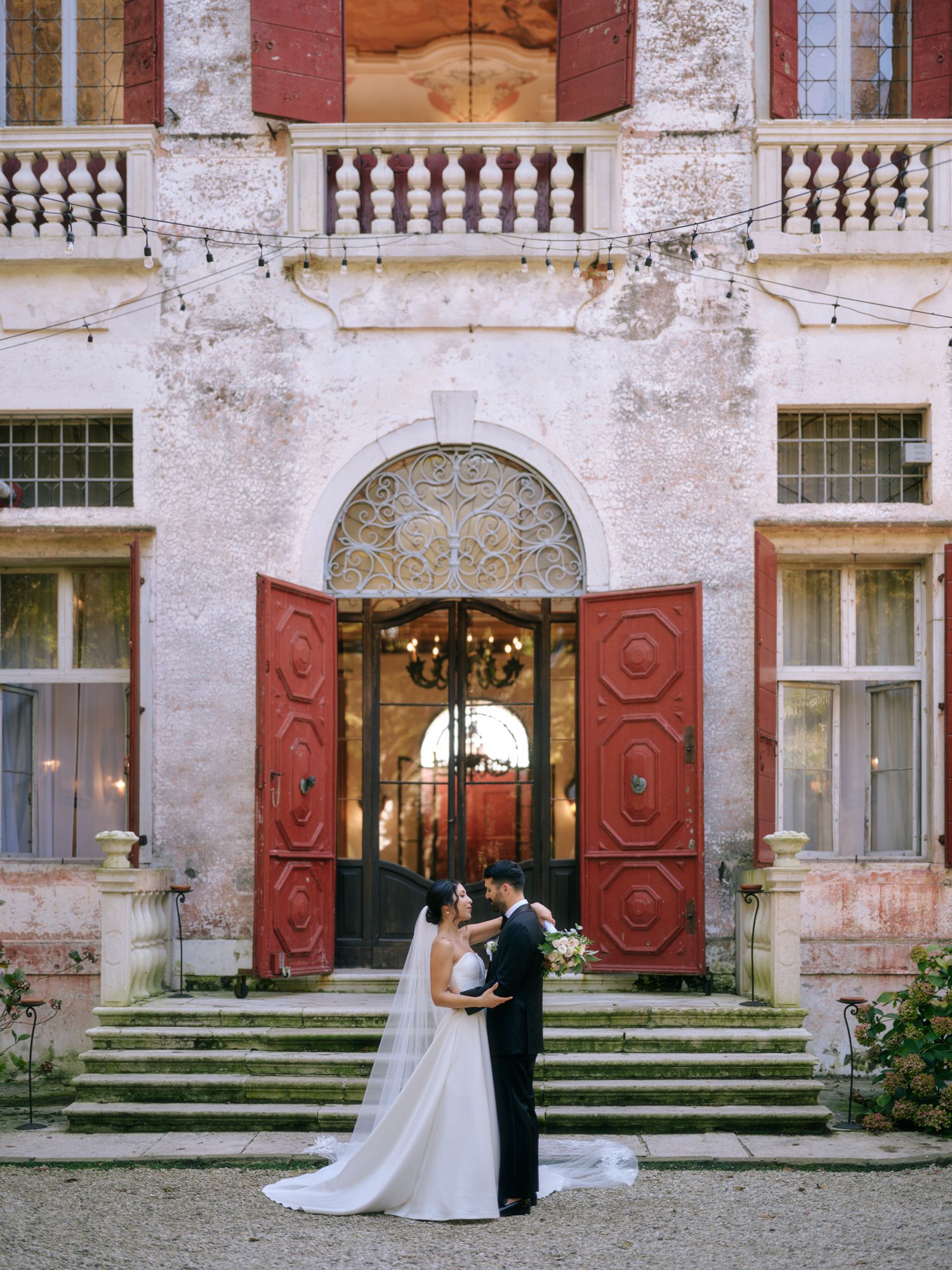A couple shares a romantic moment outside a historic building with red shutters and stone steps.