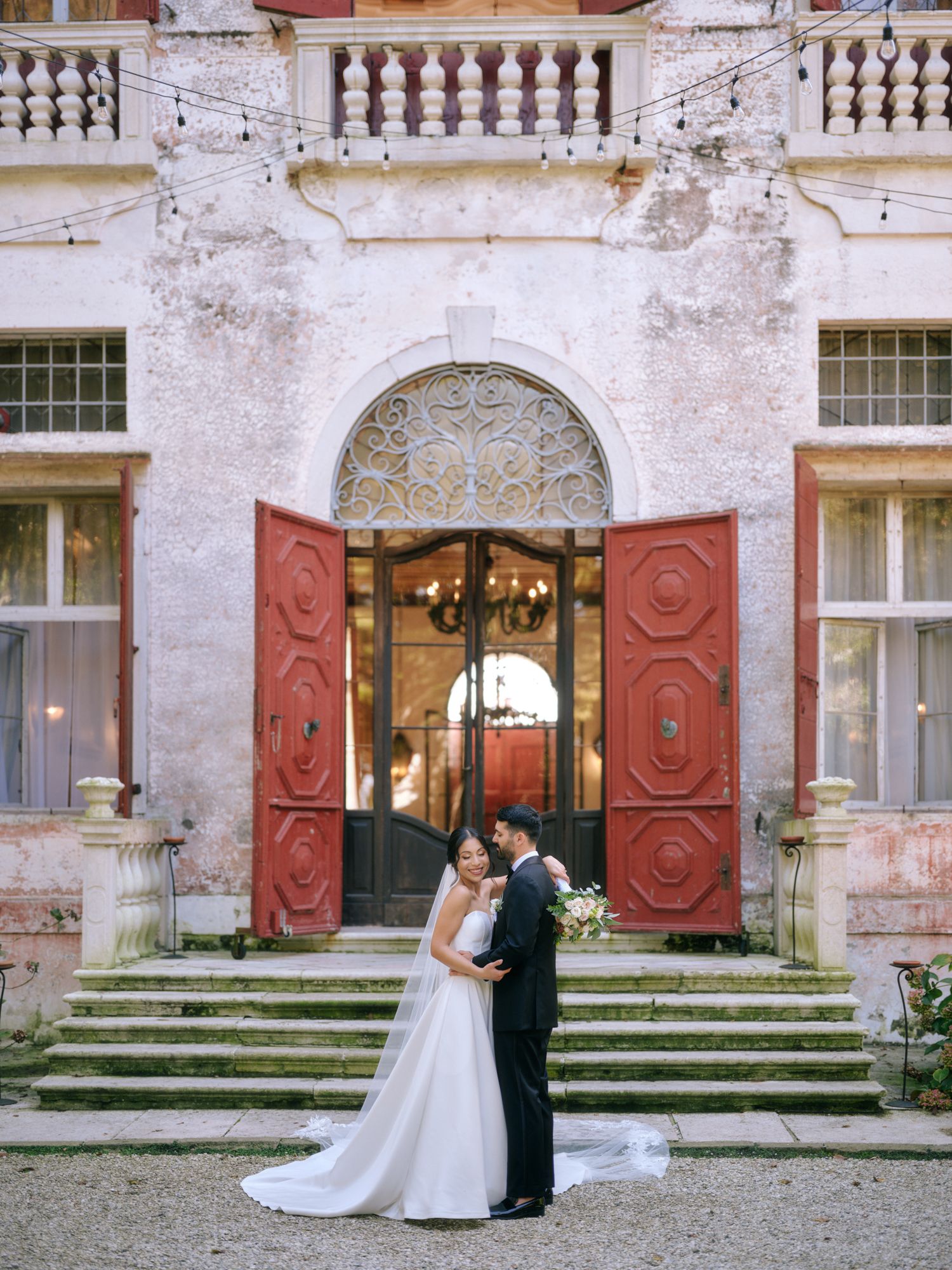 Elegant wedding couple poses in front of a historic building with red doors and stone steps in a romantic outdoor setting.
