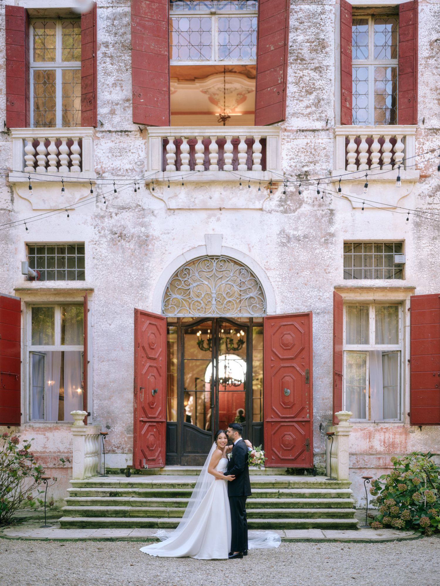 A couple stands together on the steps of a weathered mansion with red doors and ornate architecture.