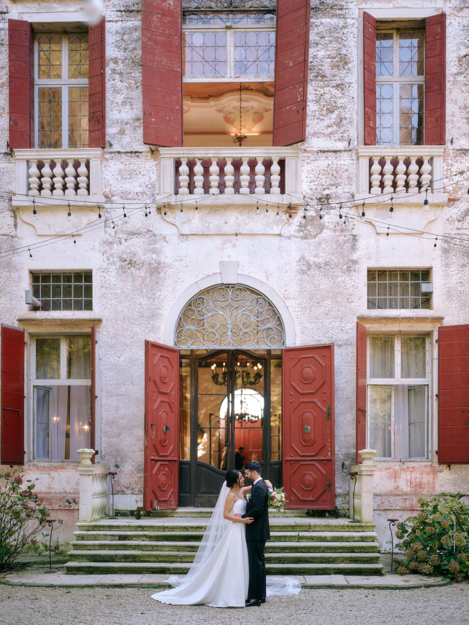 A couple poses in front of a grand historic building with red accents and stone balustrades.