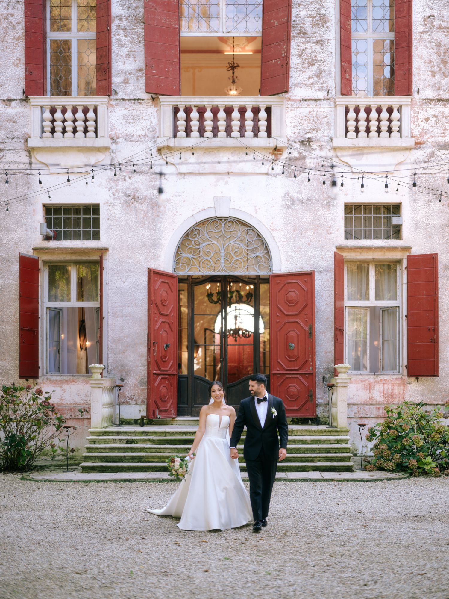 A couple walks along a grand historic villa's courtyard with ornate red and white architectural details and stone balustrades.
