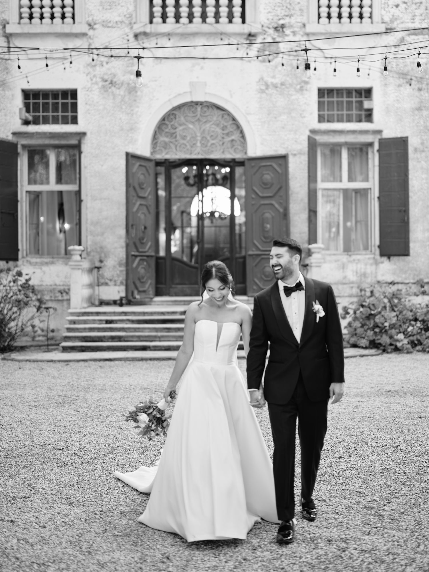 A couple walks together across a courtyard in front of an elegant European-style mansion.