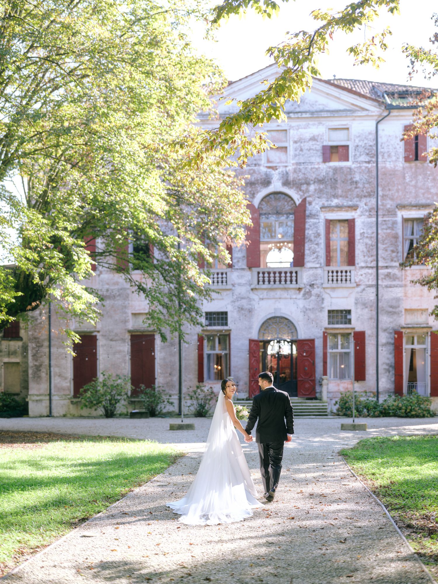 A couple walks along a tree-lined path towards an elegant historic mansion with stone facade and arched windows.