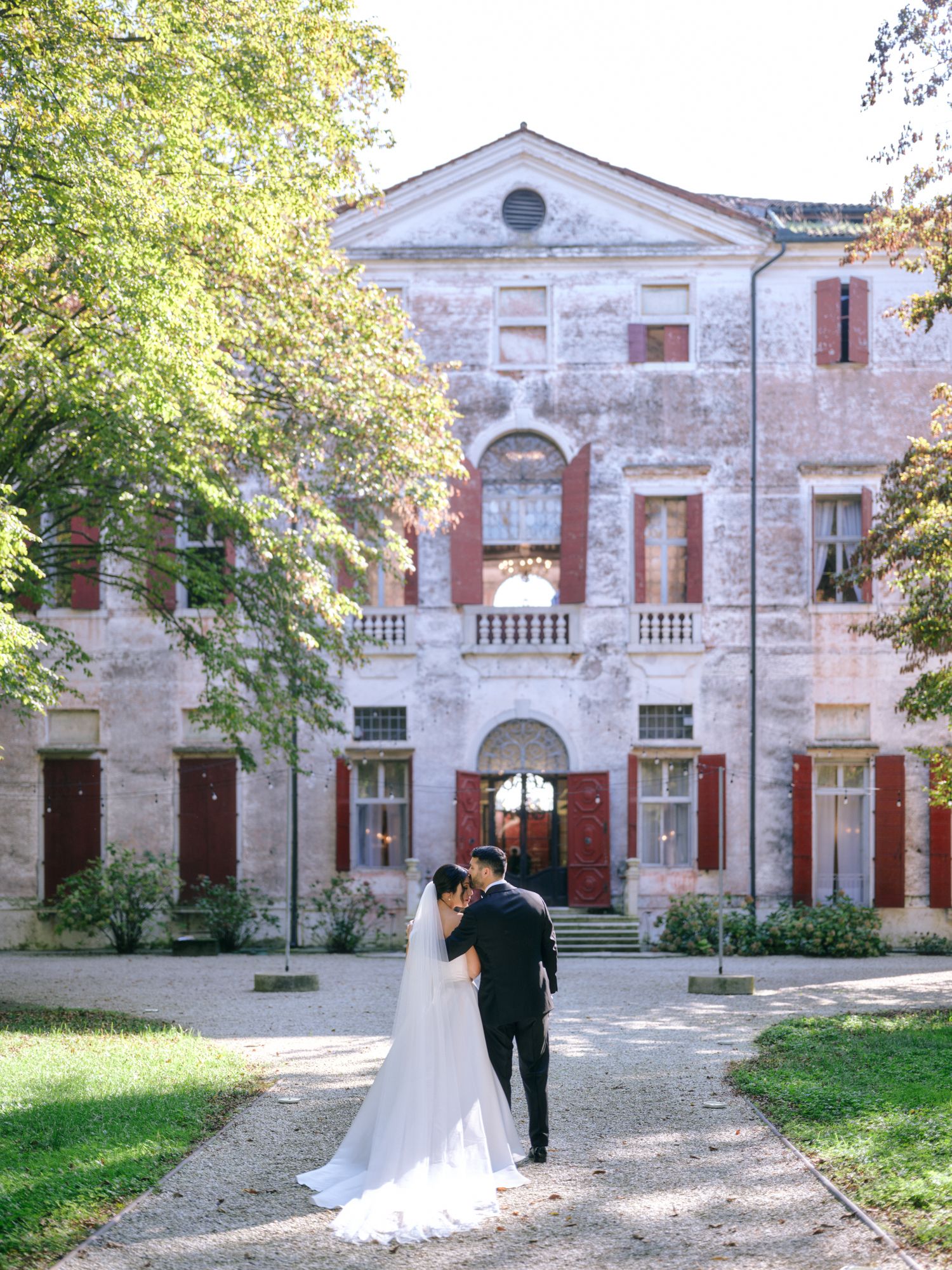 A wedding couple walks along a historic villa facade with red brick and white stone architectural details in a romantic setting.