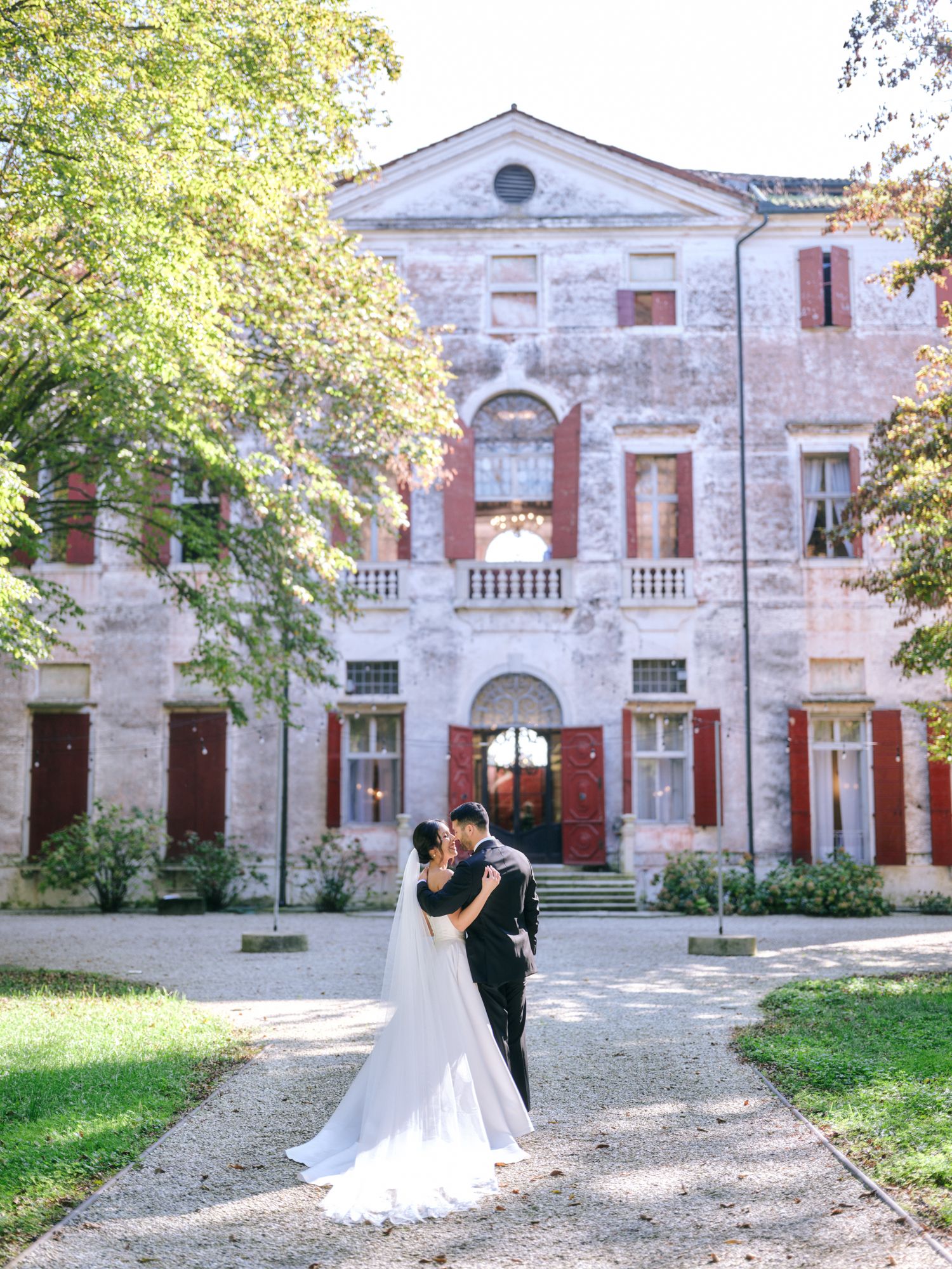 A romantic wedding couple embraces in front of a historic stone mansion with elegant architecture and green landscaping.