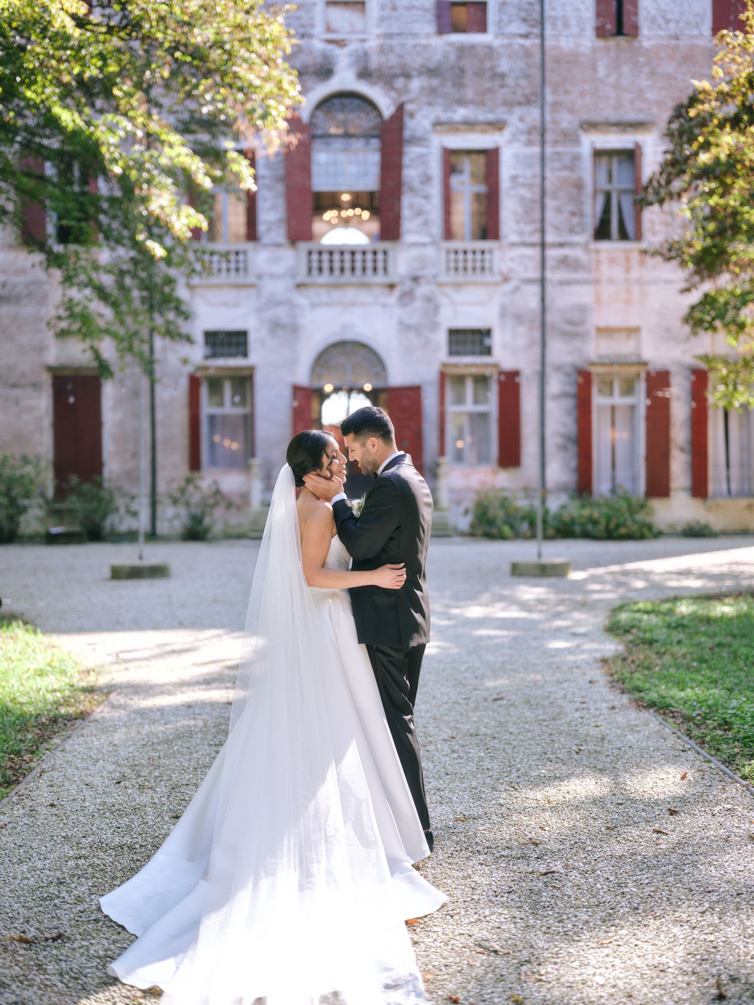 A couple in wedding attire embraces in a romantic outdoor photoshoot at a historic stone building courtyard.