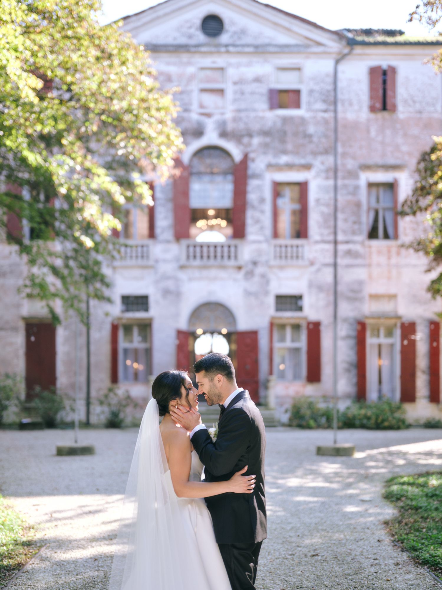 A romantic wedding couple embraces in front of a grand historic stone mansion with red shutters and elegant architecture.