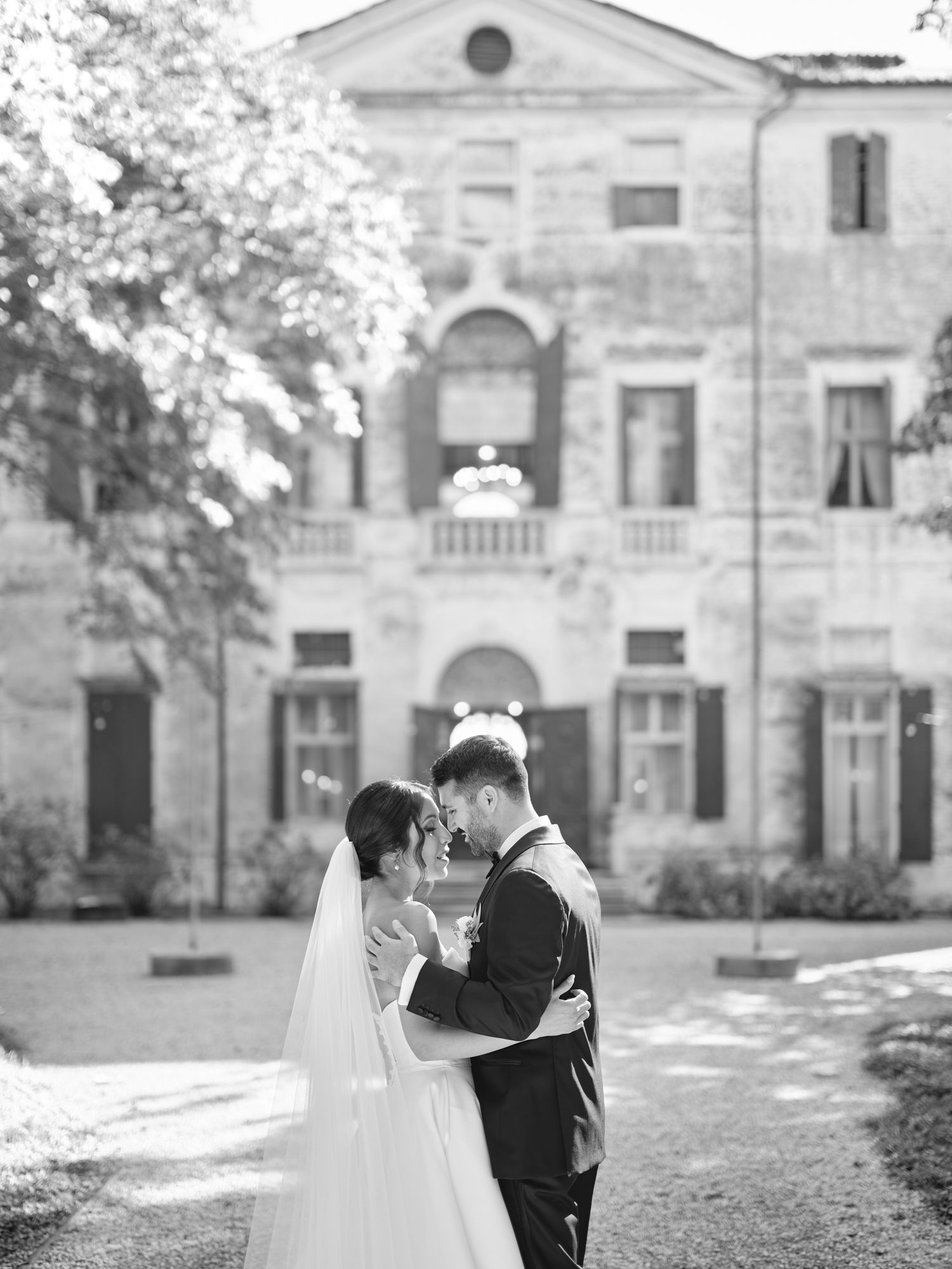 A romantic black and white wedding photo shows a couple embracing in front of a grand historic mansion.
