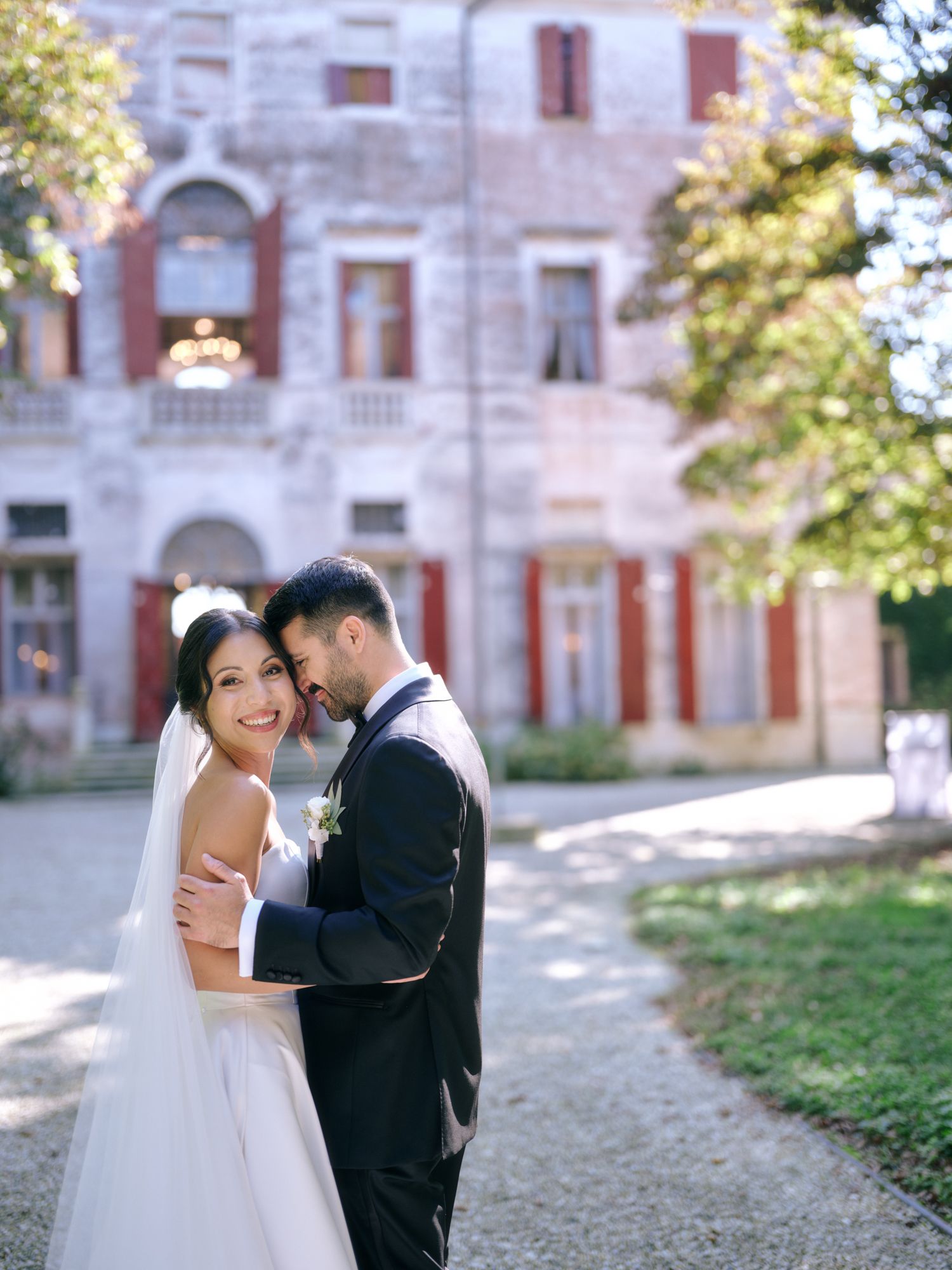 A romantic wedding couple embraces on a path in front of a grand historic mansion with arched windows.