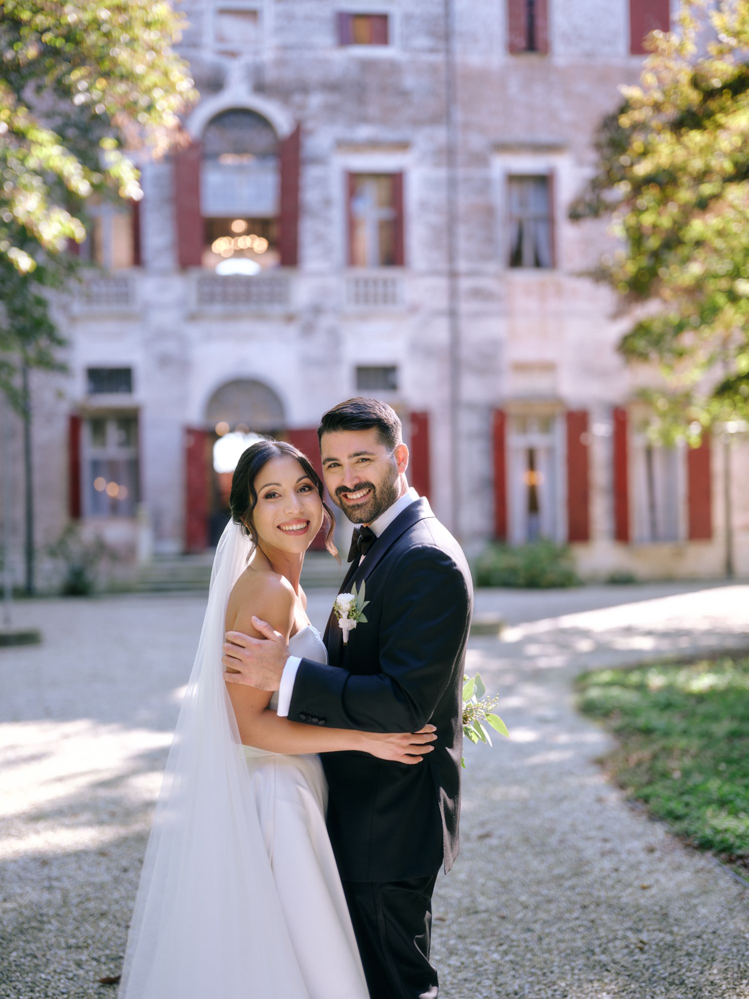 A couple in wedding attire embraces and laughs together on the grounds of a historic stone building.