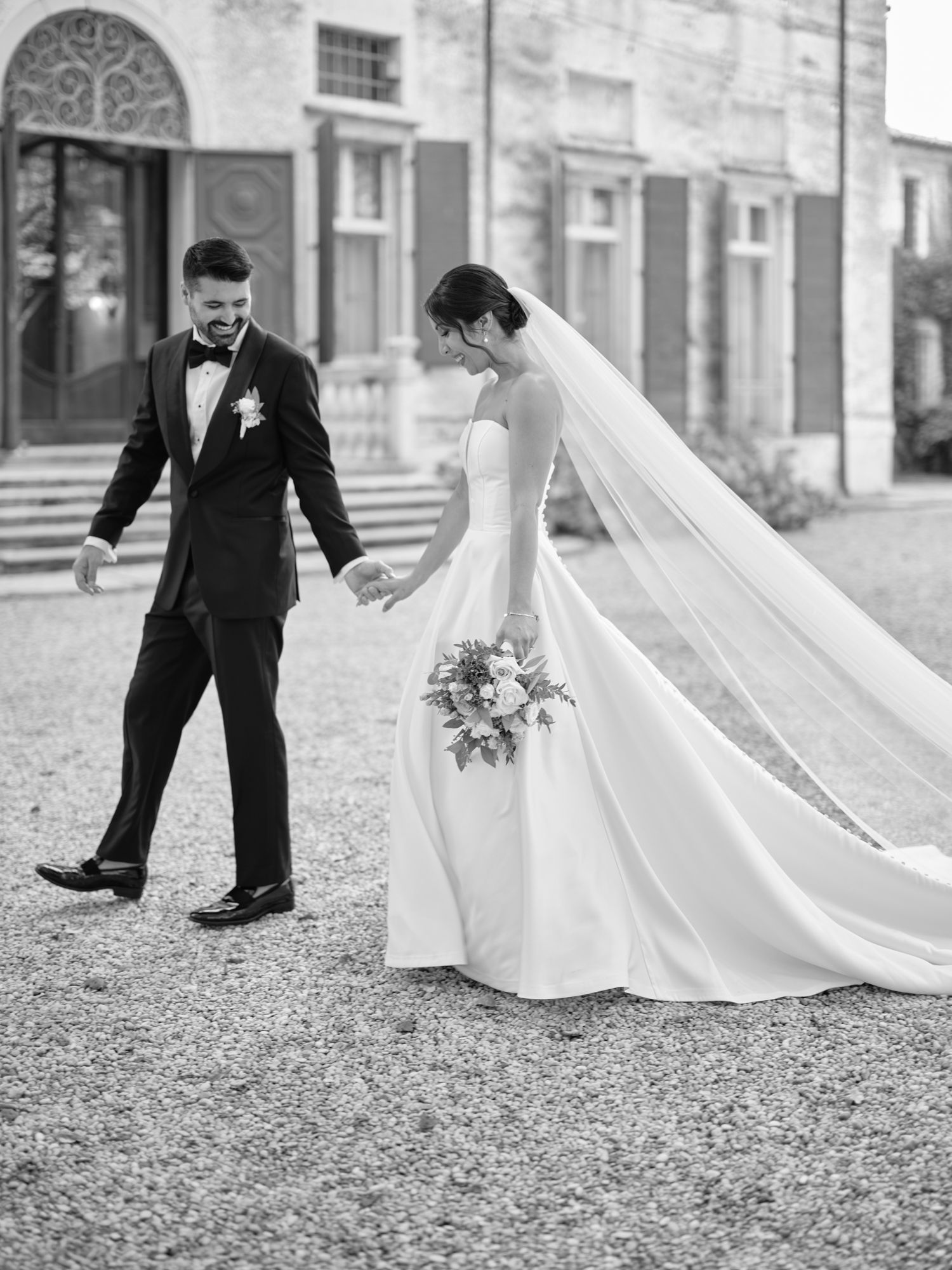 Black and white wedding photo showing a bride in flowing dress with long veil holding bouquet walking with groom in tuxedo.