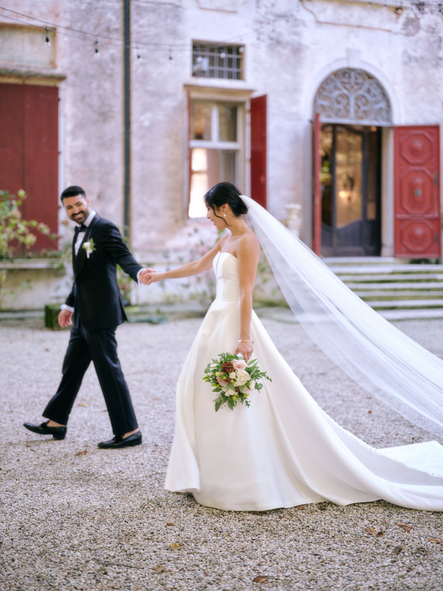 A couple in wedding attire playfully poses on a gravel courtyard outside a historic building with red doors.