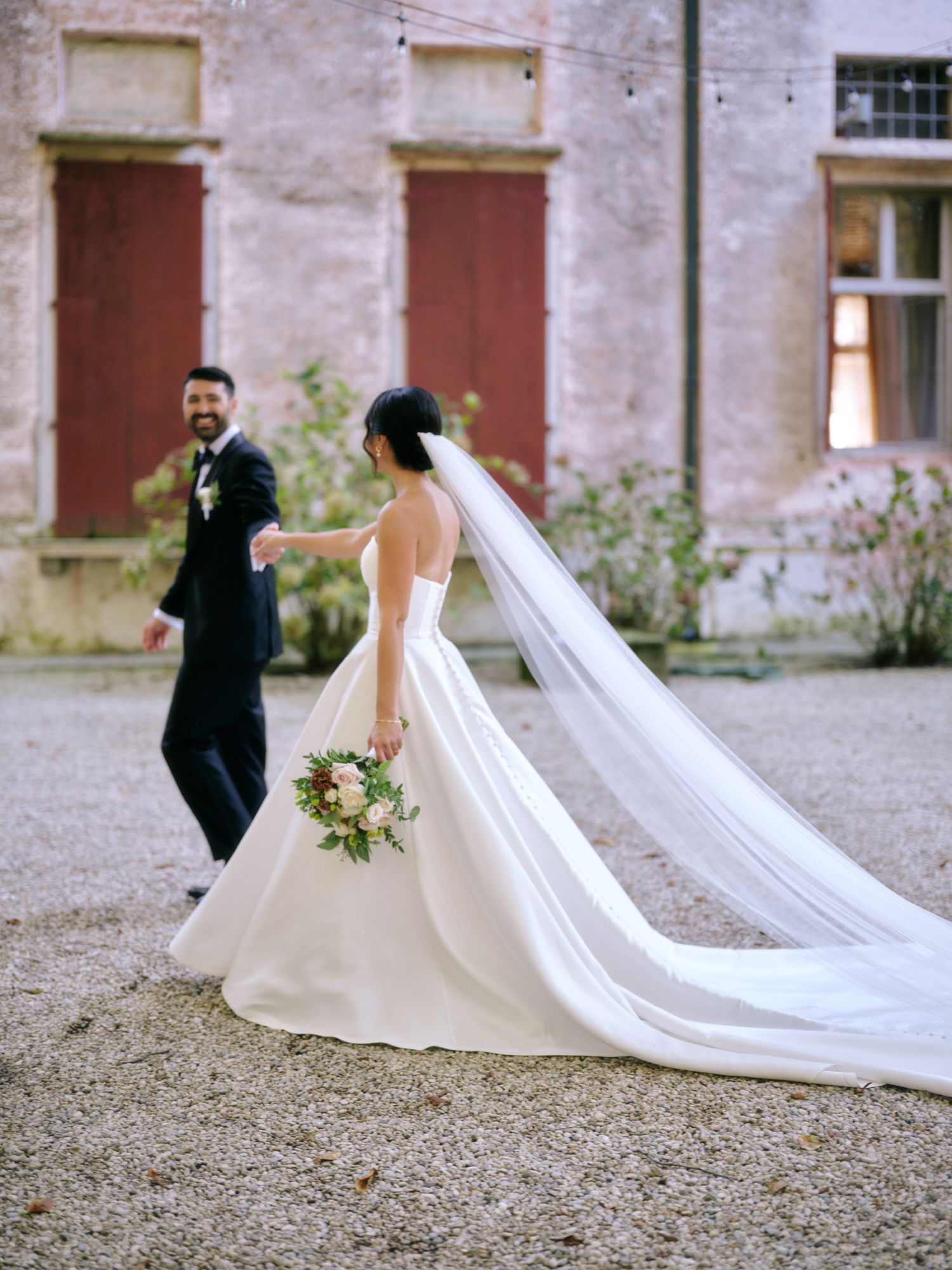 A wedding couple in formal attire walks outside a rustic building, with the bride's long cathedral veil trailing behind.
