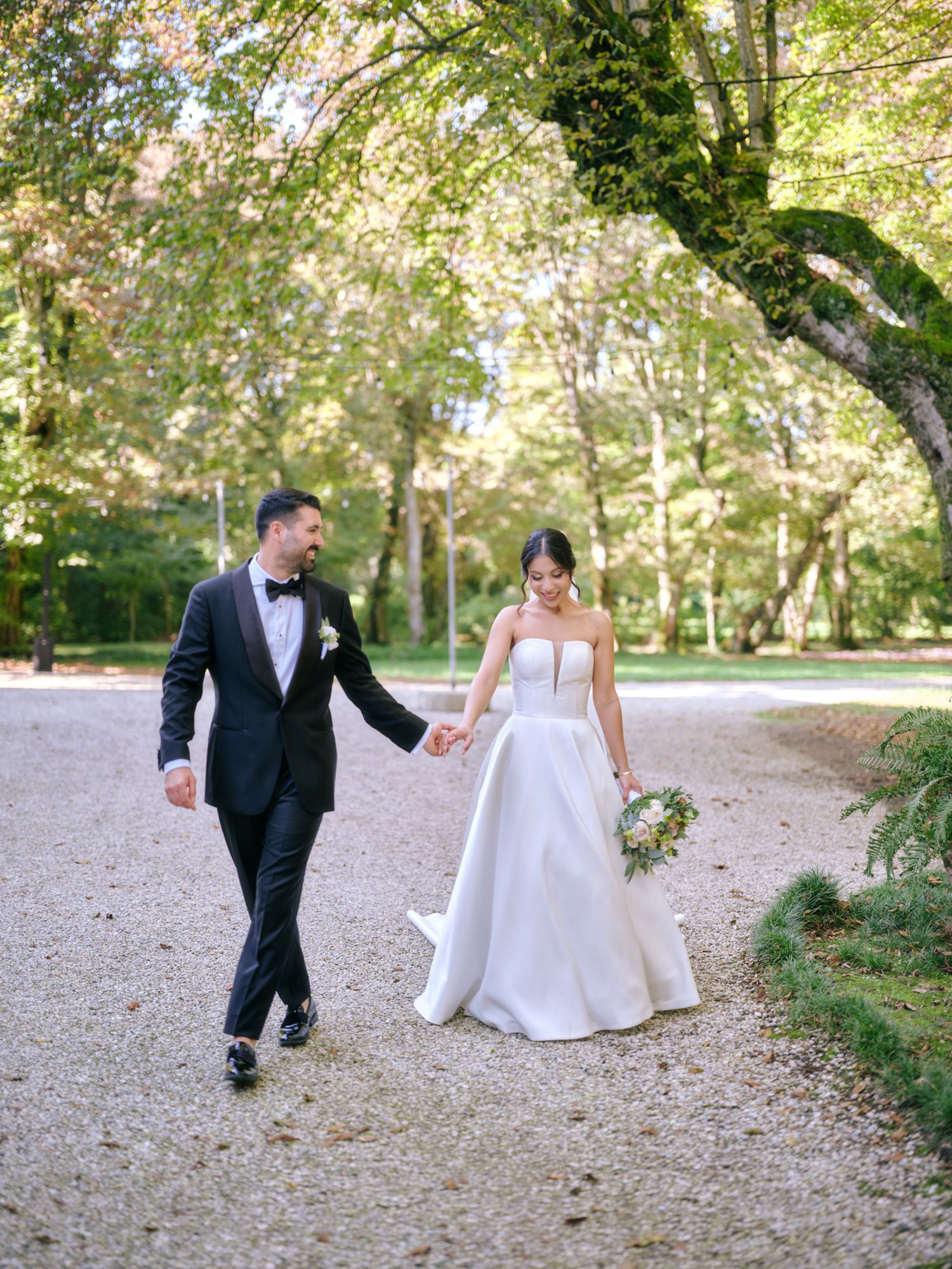 A couple in formal wedding attire walk together on a leaf-covered path through a wooded park setting.