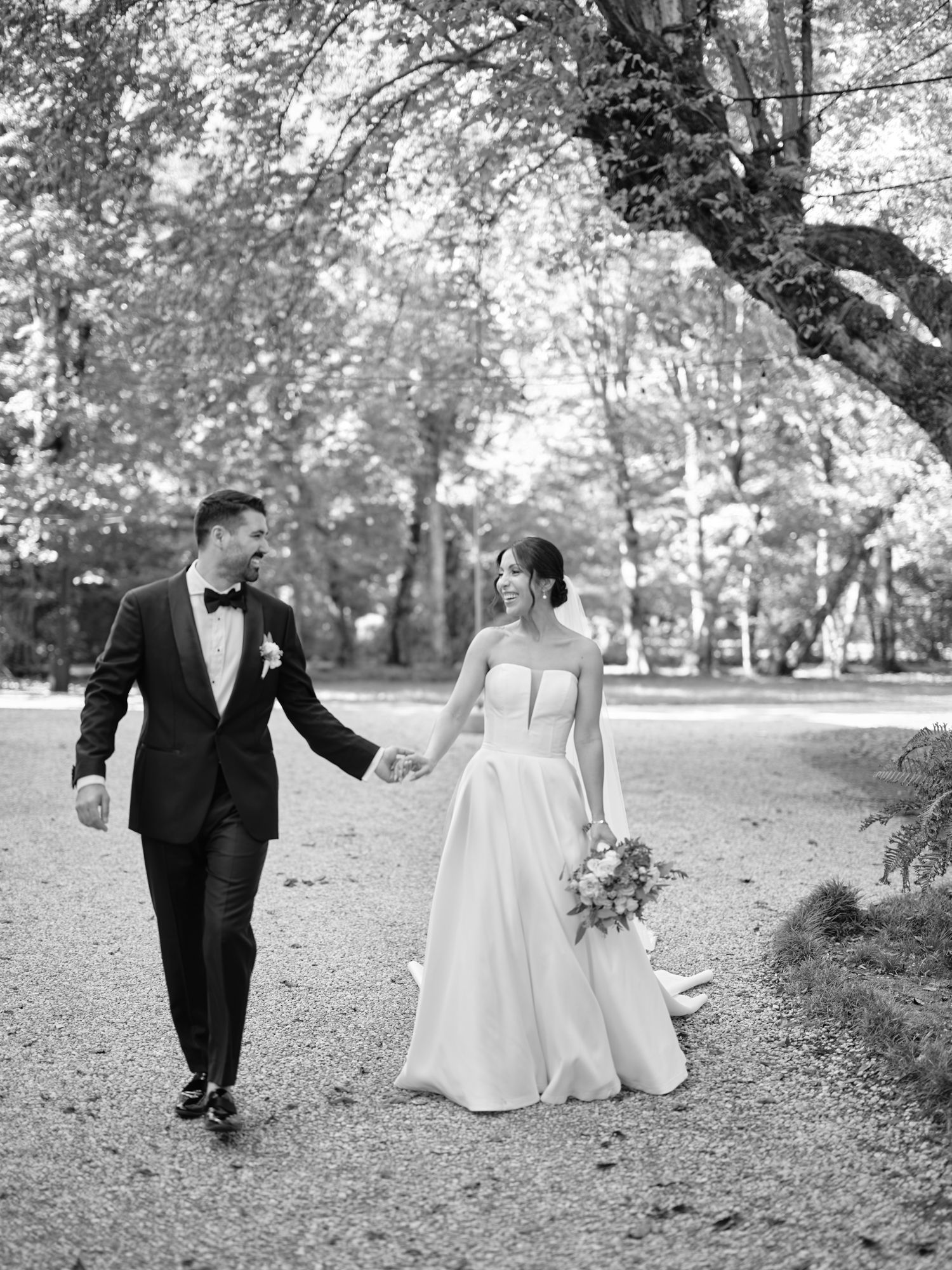 A bride and groom walking together on a tree-lined path in black and white photography.