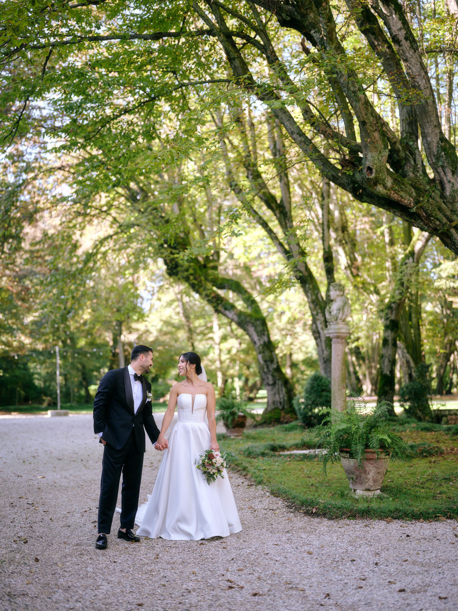 A couple walks together on a gravel path under towering trees in a romantic garden setting.