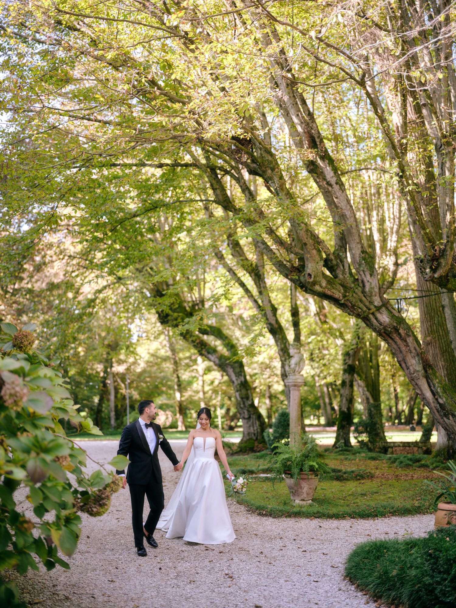 A couple walks along a scenic tree-lined path in a lush garden setting on their wedding day.