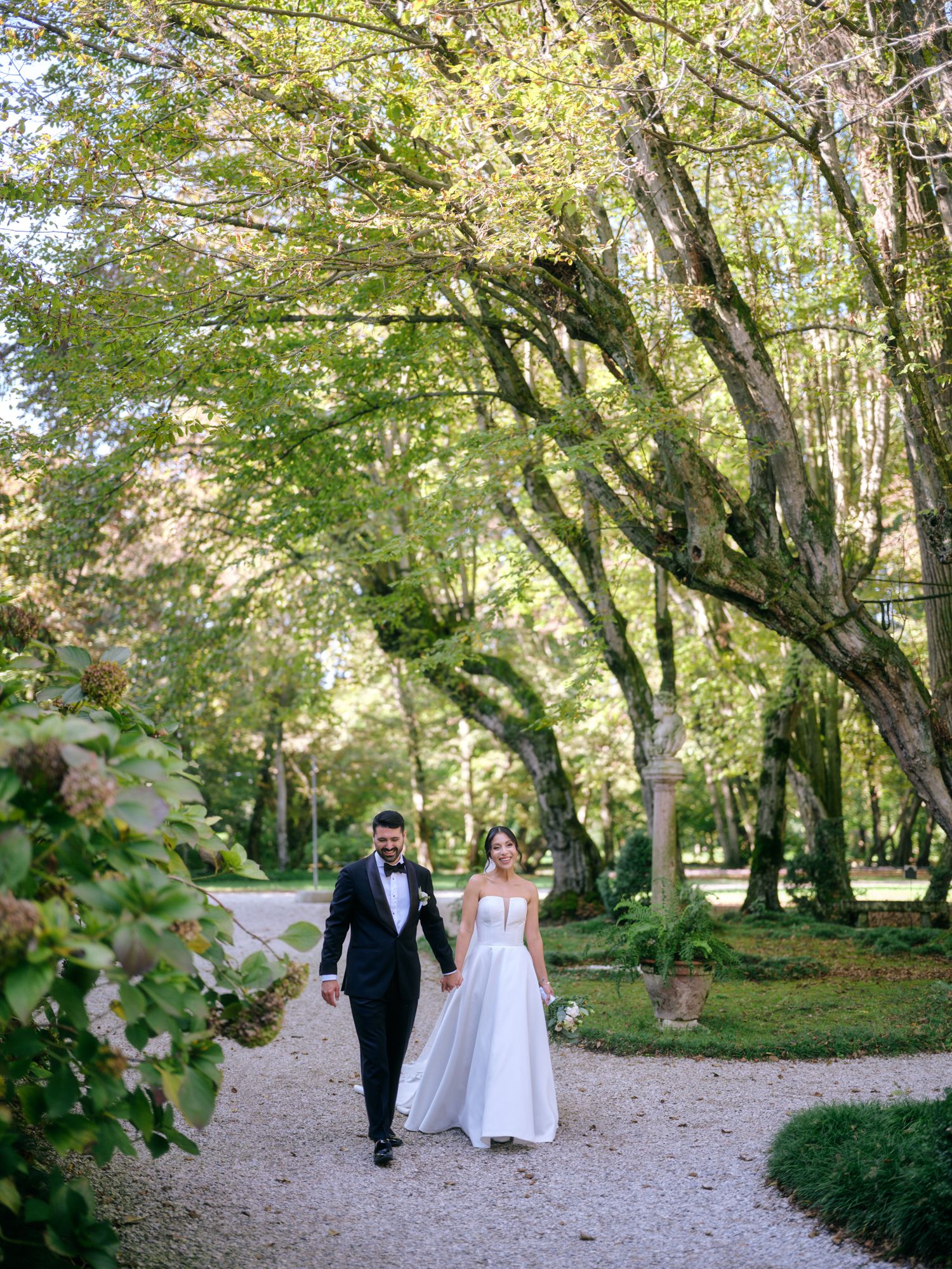 Newlyweds stroll down a winding gravel path surrounded by green trees and natural scenery.