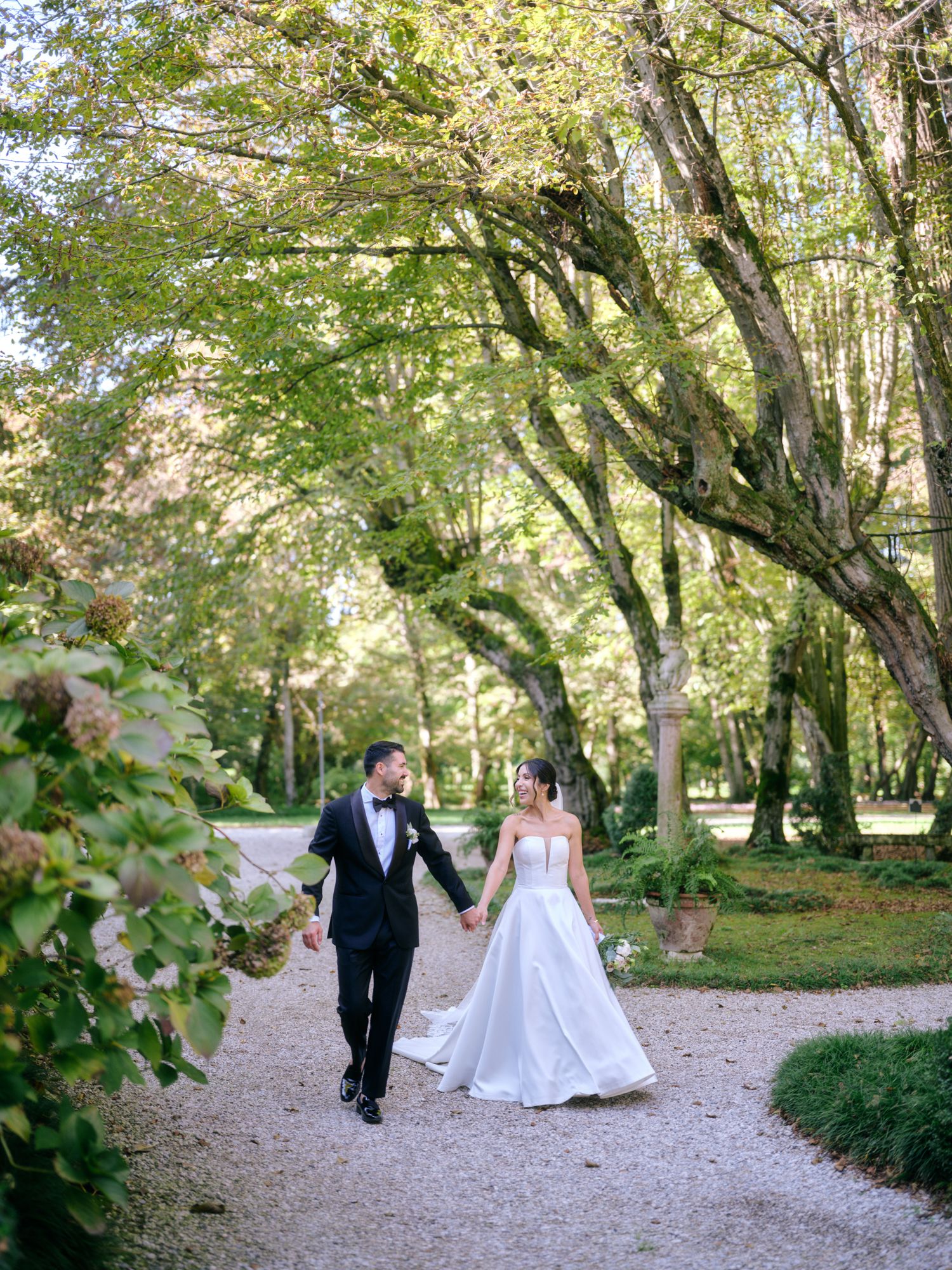 A couple walks hand in hand along a serene garden path surrounded by lush green trees and foliage.