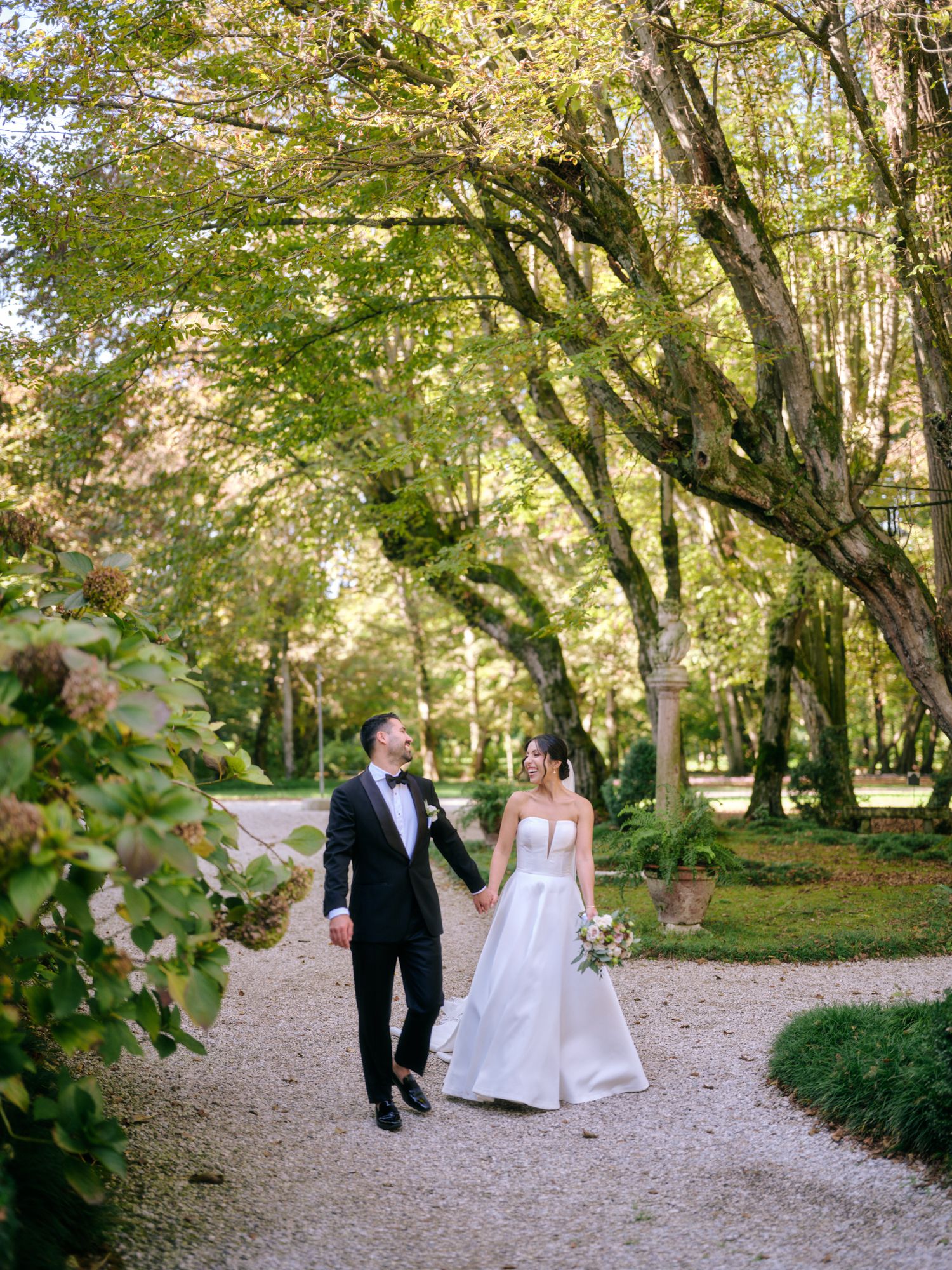 A couple walks hand in hand down a gravel path surrounded by lush green trees in a romantic garden setting.