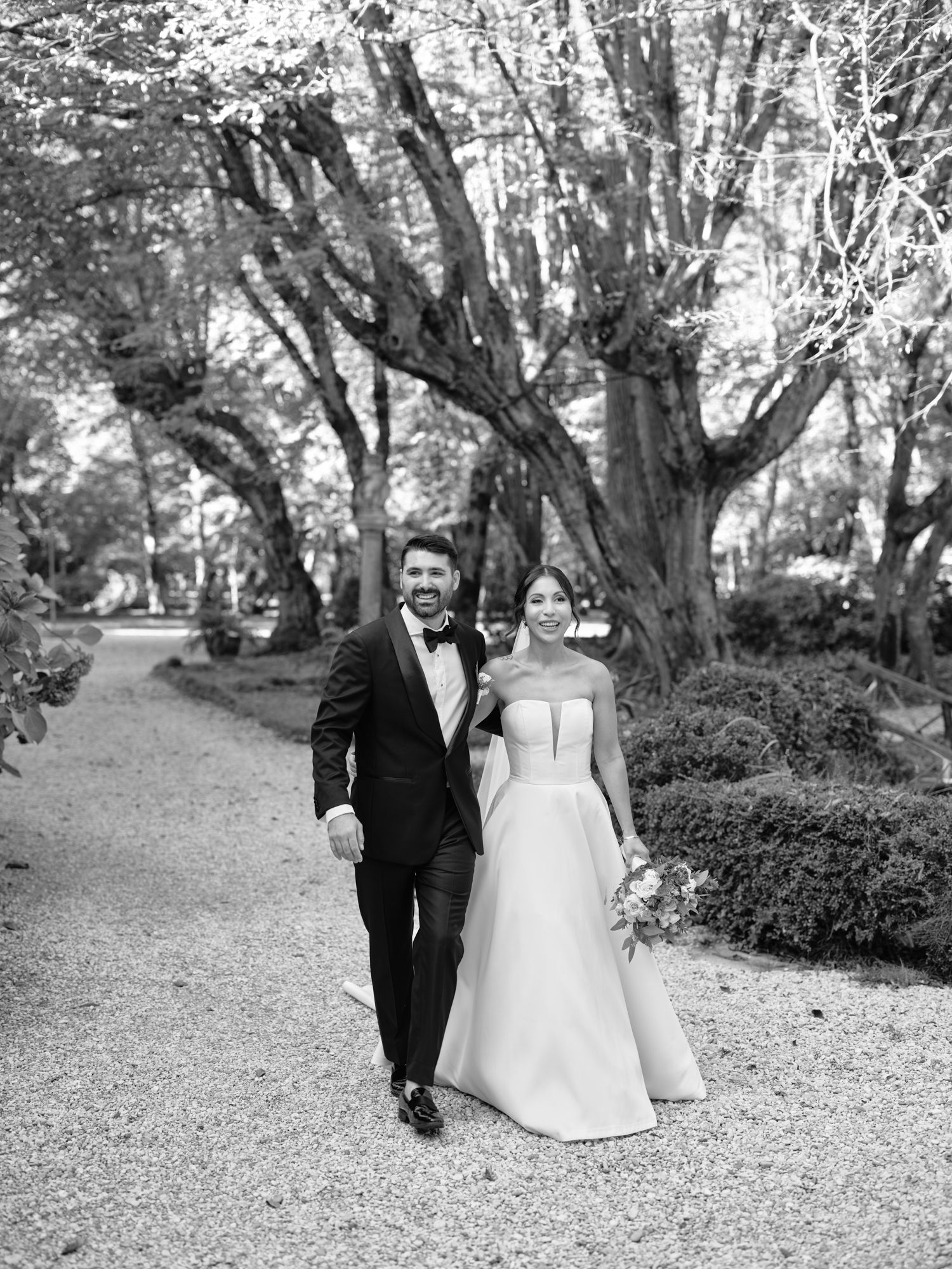 Black and white photo of a wedding couple walking together on a gravel path through a wooded garden.