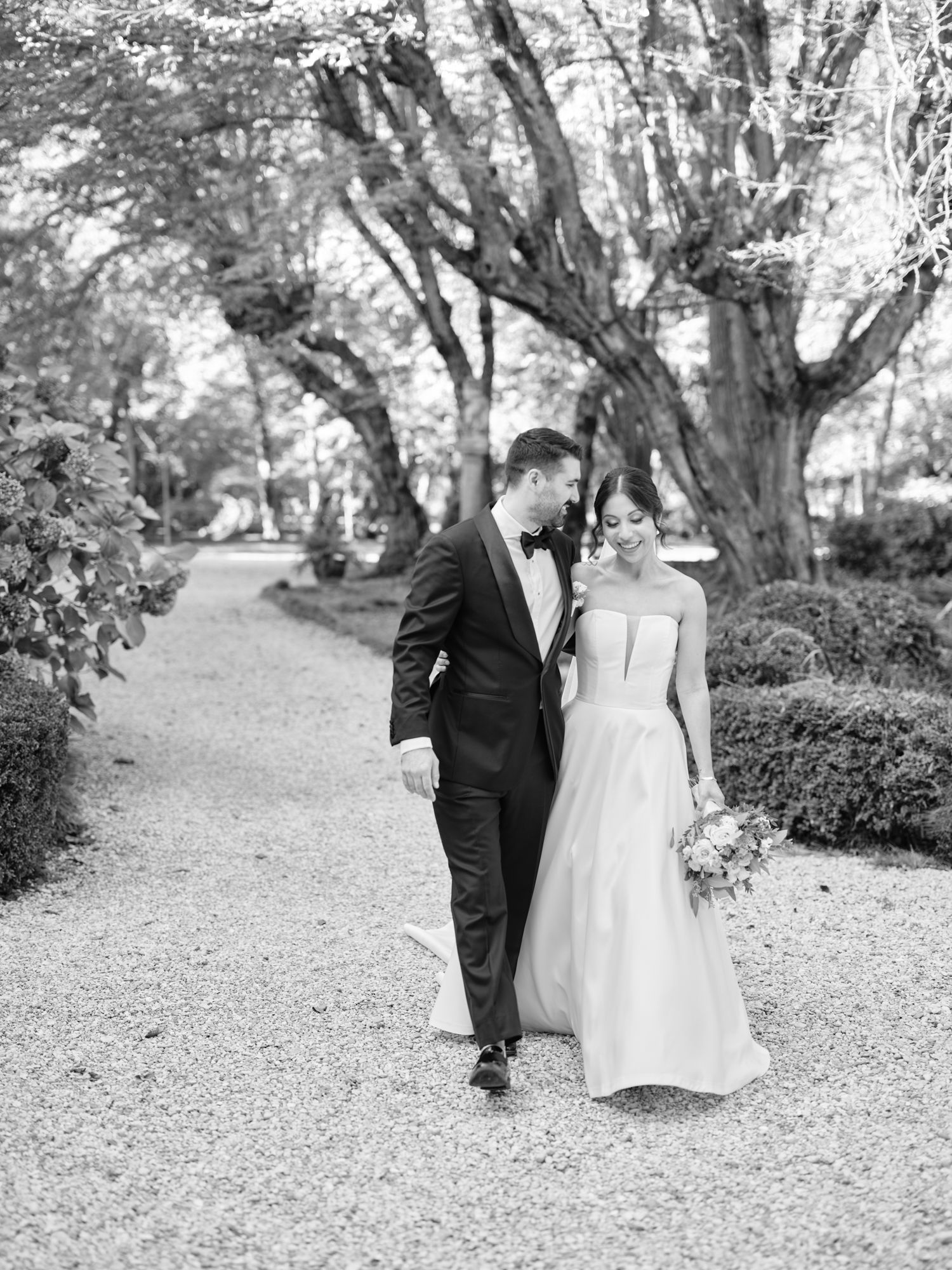 A couple in wedding attire walking down a tree-lined garden path in black and white photography.