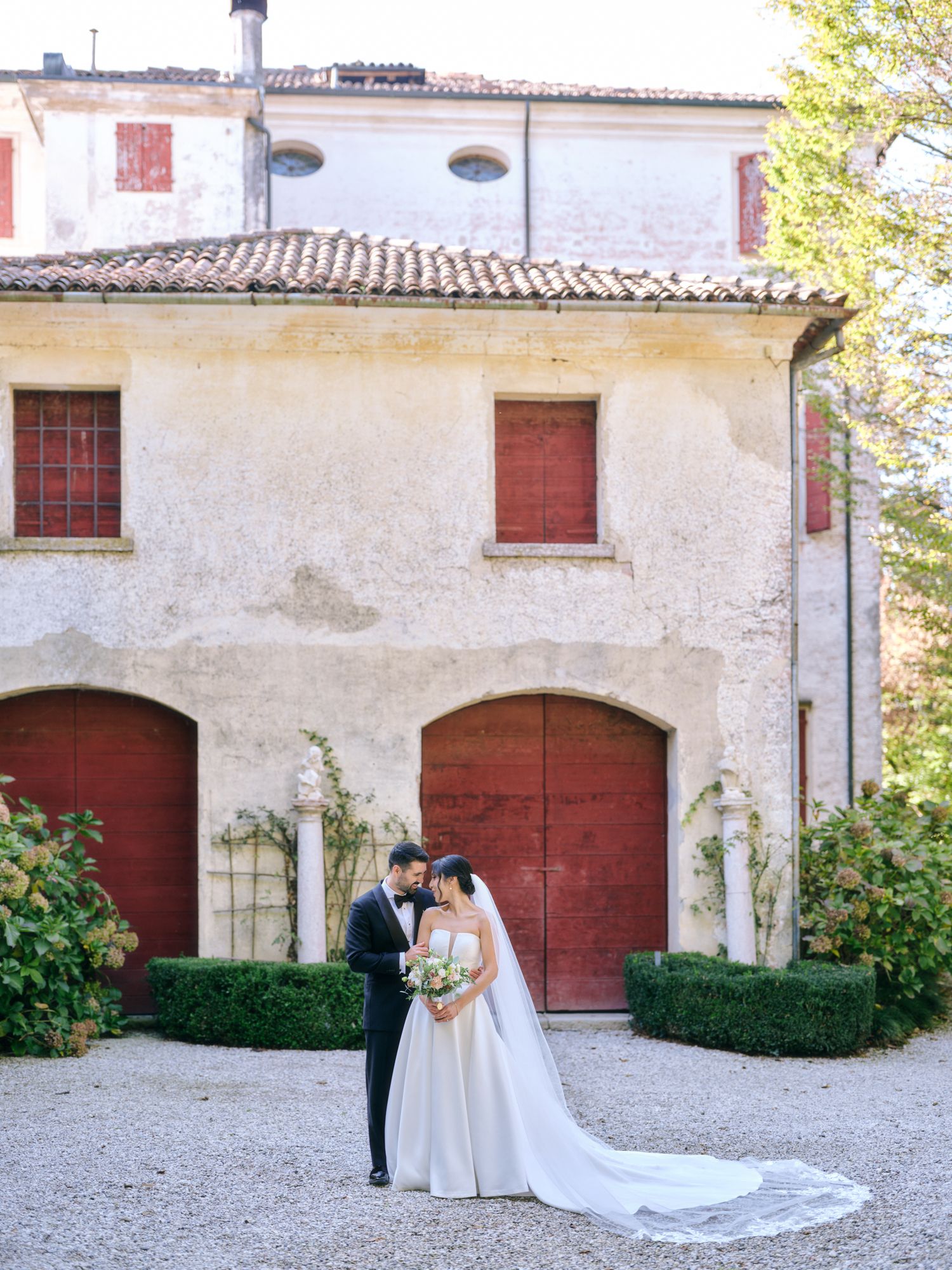 A romantic wedding scene captured in a rustic Italian courtyard with red doors and white stone walls.