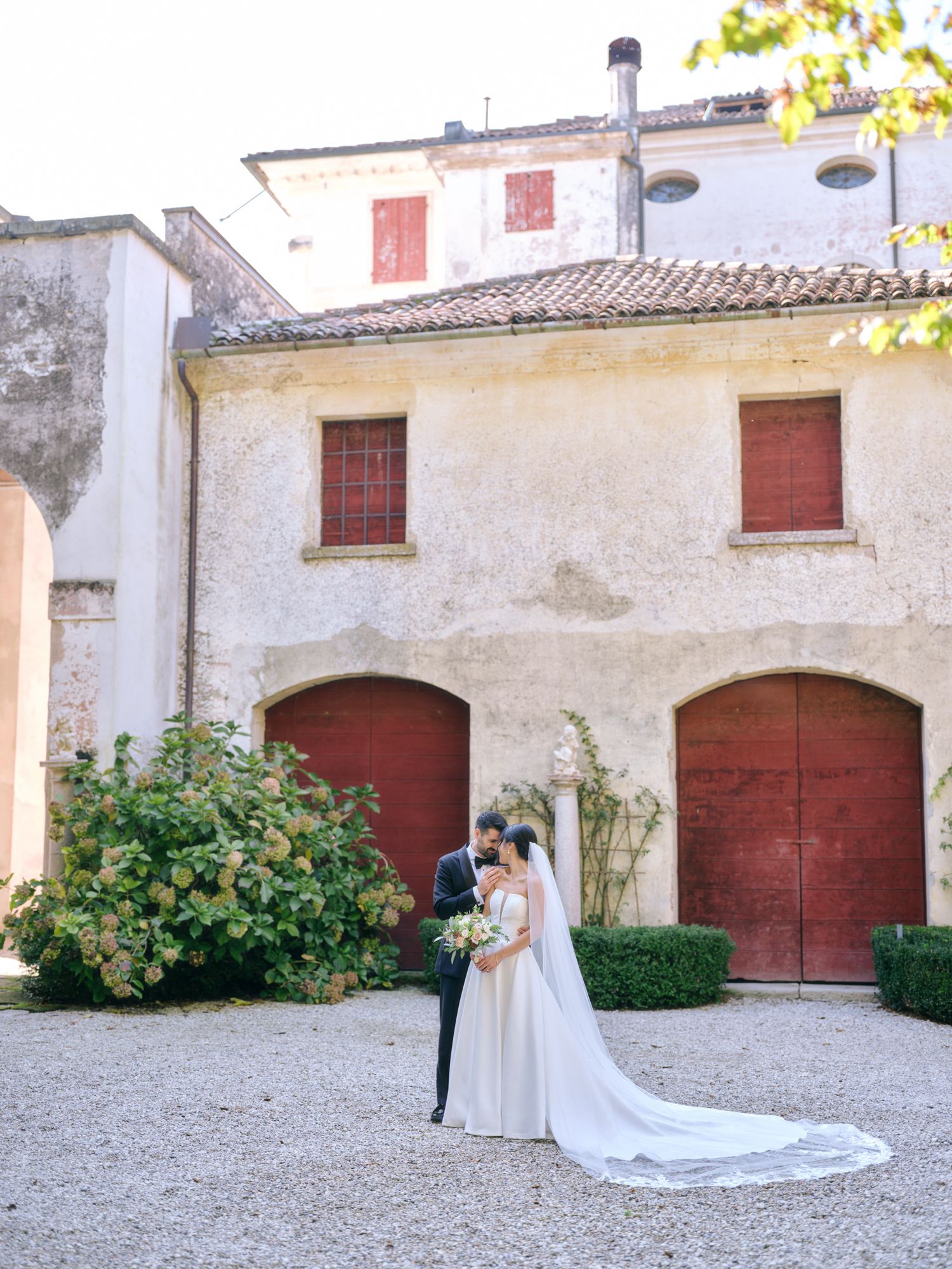 Romantic wedding couple poses against rustic Italian villa walls with long flowing white veil trailing behind.