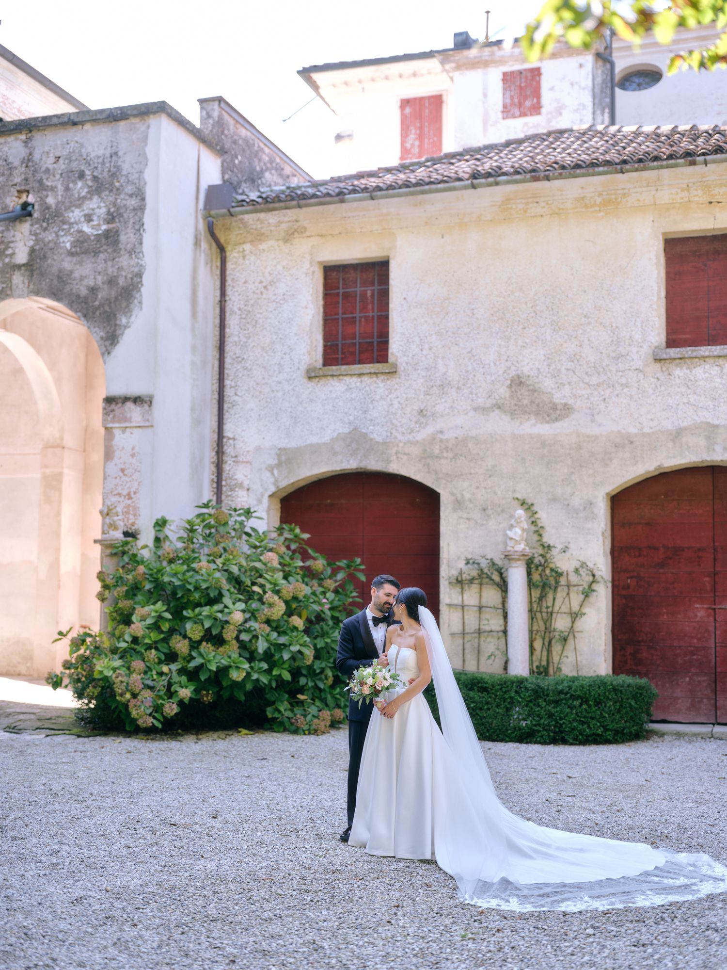Elegant couple in wedding attire stands near historic stone building with red doors and green shrubbery.