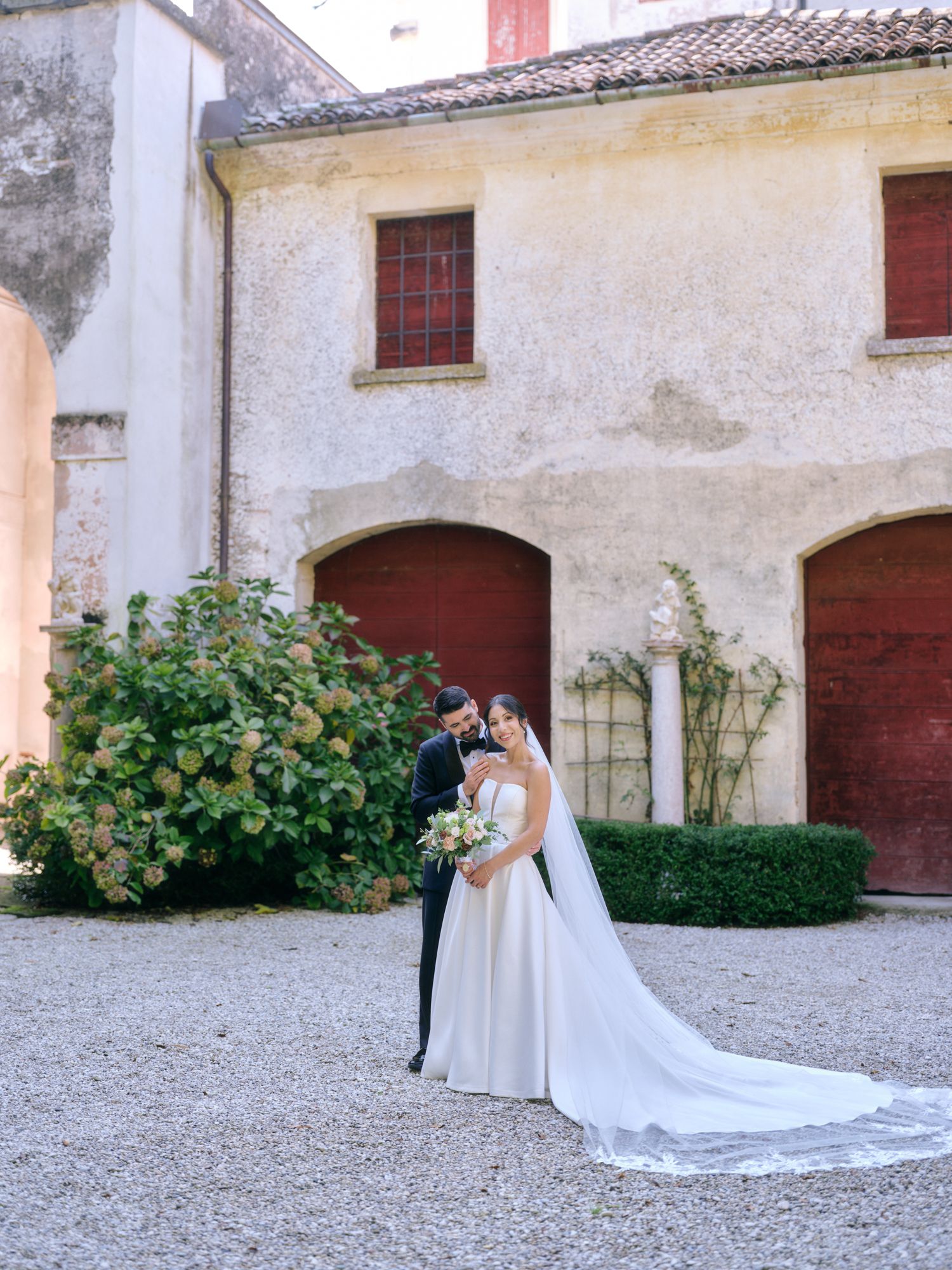 Two people in elegant wedding attire embrace in a romantic moment against a rustic stone building courtyard.