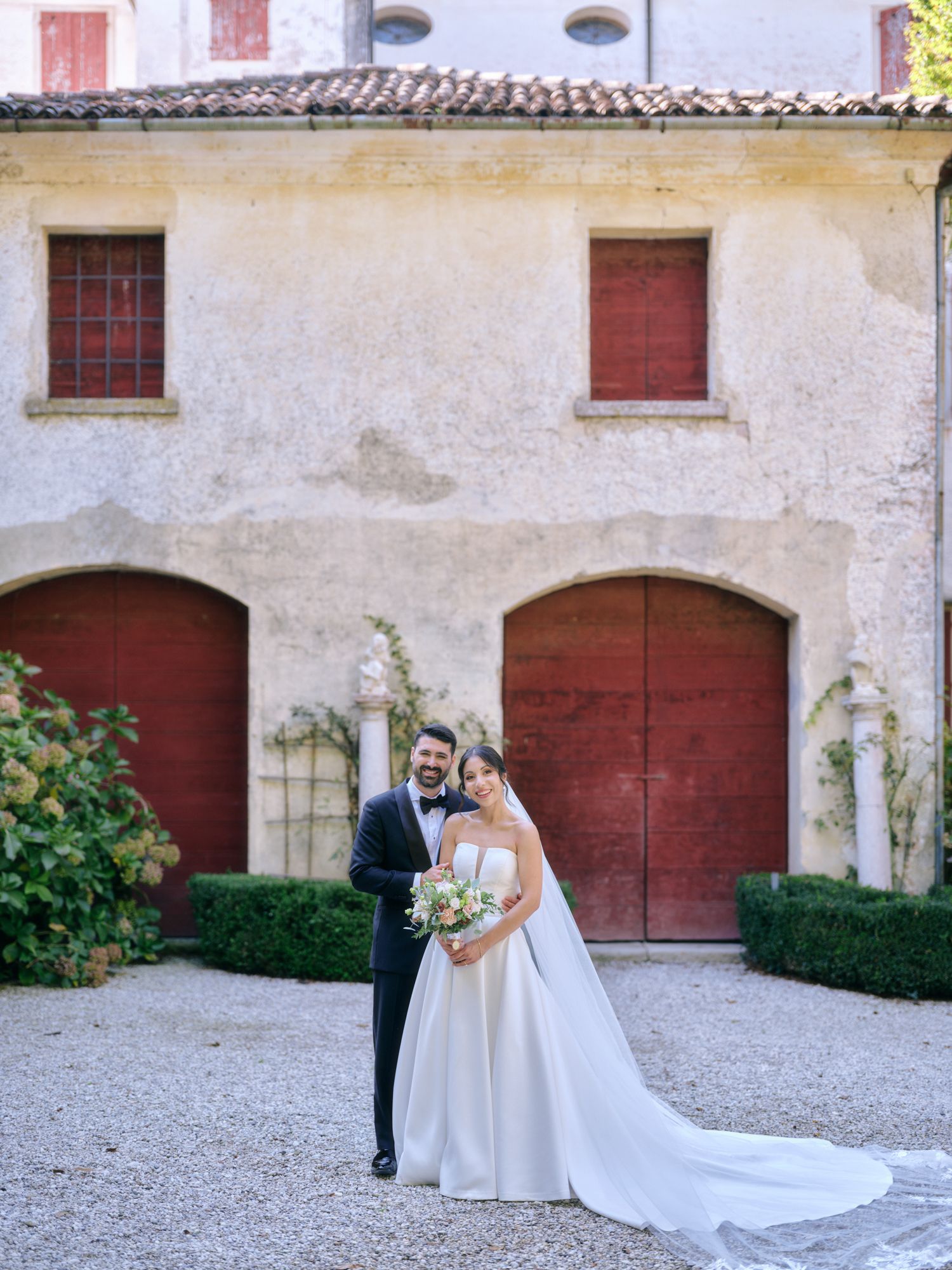 A couple stands in front of a rustic Italian villa with red doors and a tiled roof on their wedding day.
