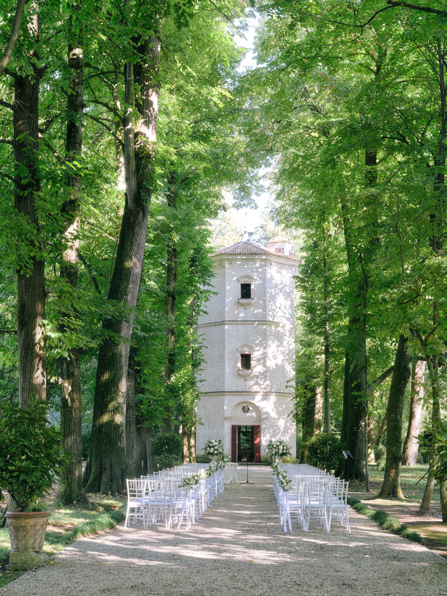White tower chapel surrounded by lush green trees with rows of white chairs set up for an outdoor wedding ceremony.