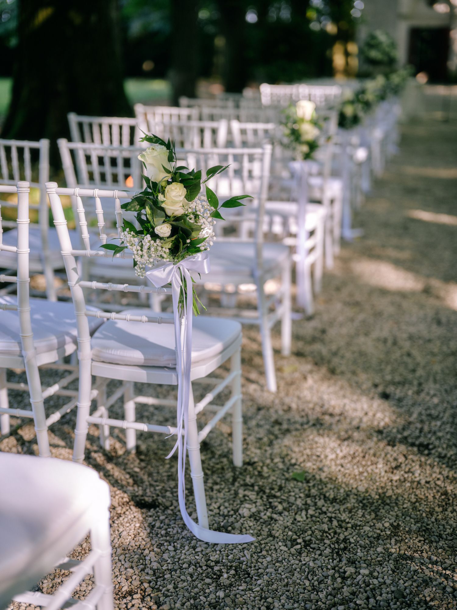 White chiavari chairs line an outdoor wedding aisle with elegant floral arrangements tied with flowing ribbons.