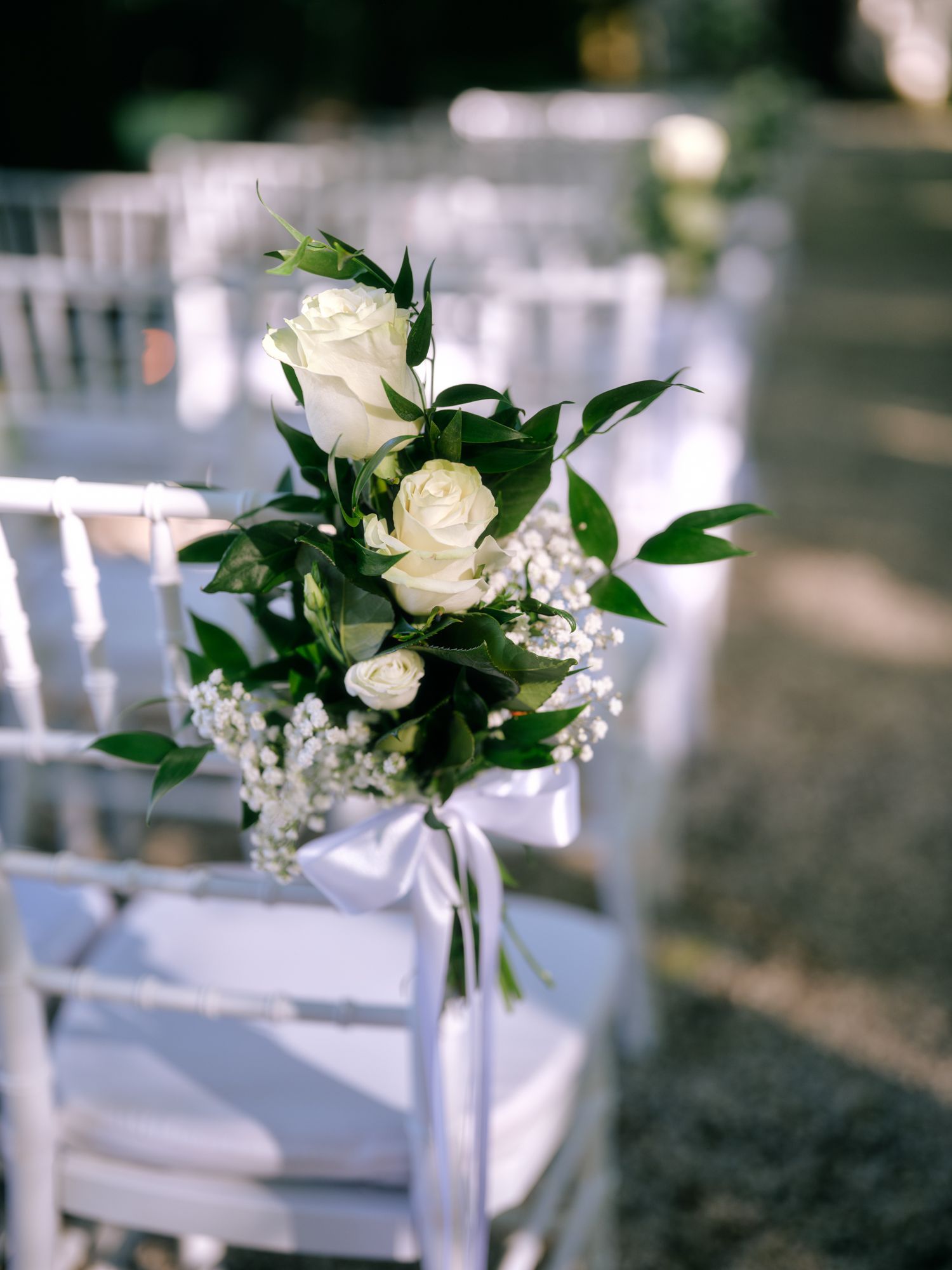 White roses and greenery floral arrangement tied with ribbon decorates a white wooden chair at an outdoor wedding ceremony.